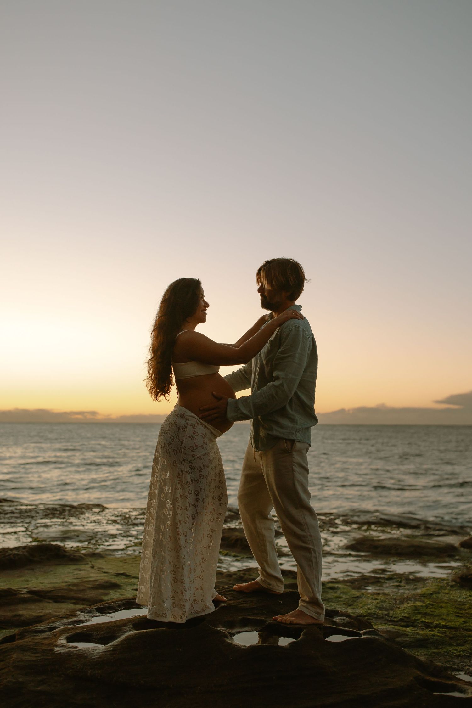 Silhouetted couple embraces on rocky shoreline during golden sunset over calm ocean waters.