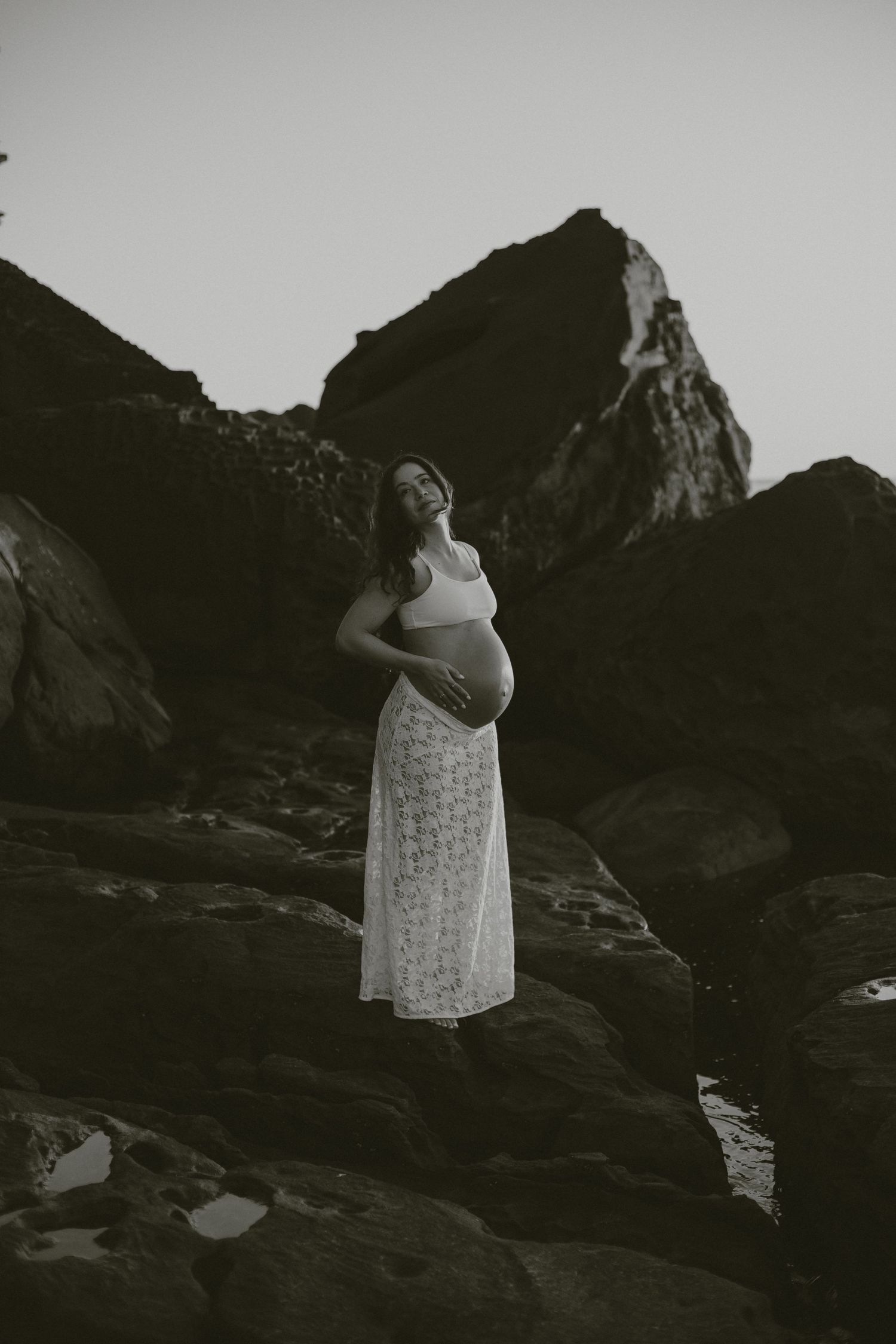 A silhouette of a pregnant figure in a lace dress stands against dramatic mountain peaks at dusk.