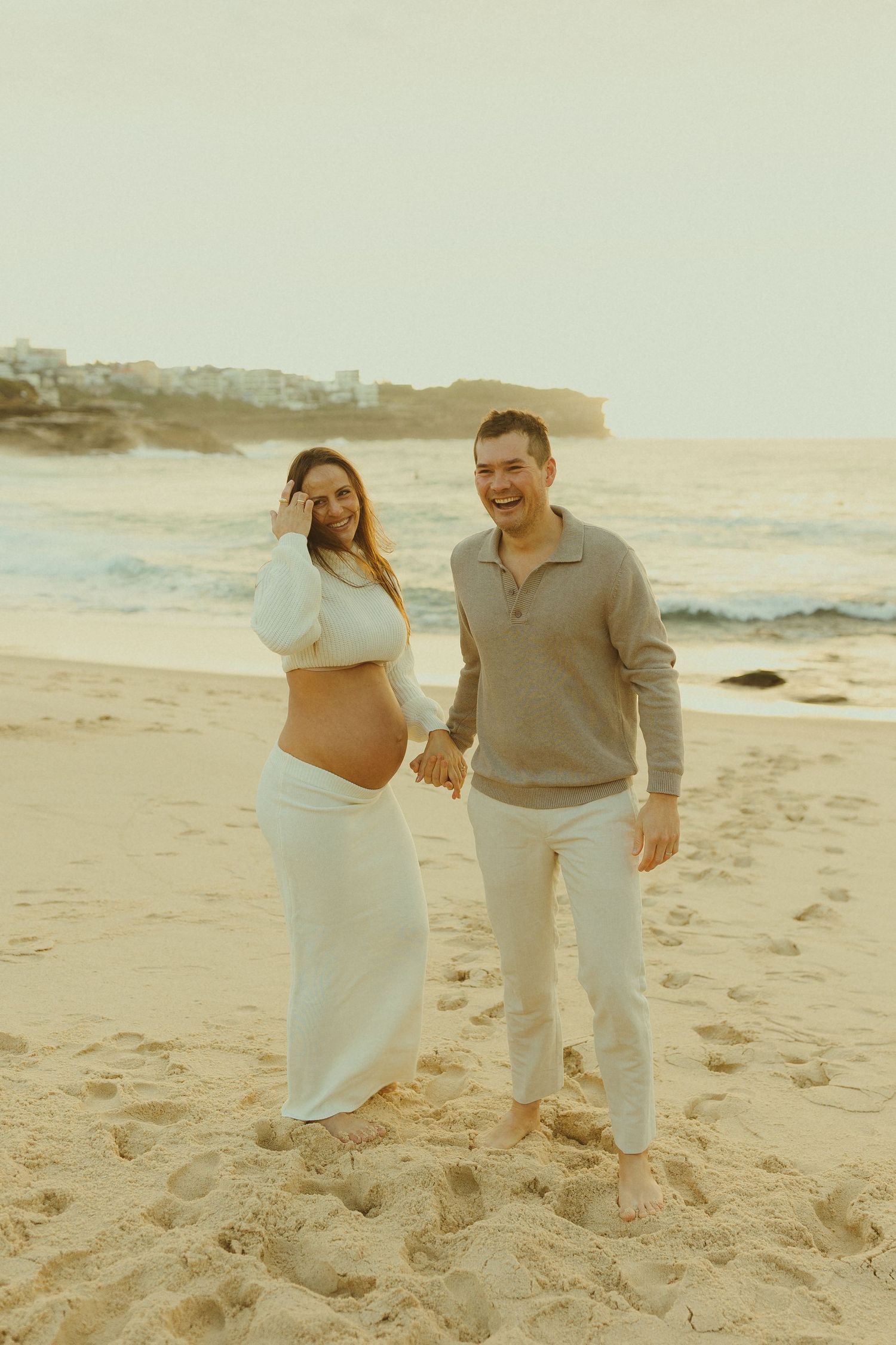 Expecting couple in white outfits share an intimate moment on a sunset beach during maternity photoshoot.