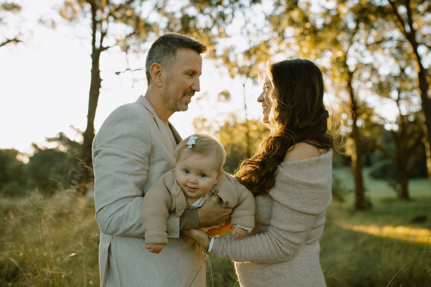 A couple in neutral-toned sweaters share a tender moment with their baby in a sunlit outdoor setting during golden hour.