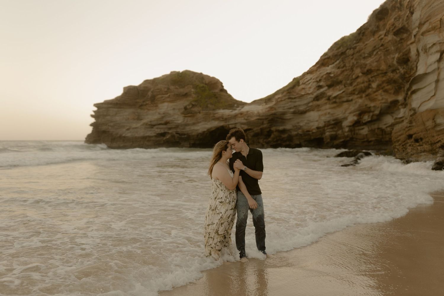 Couple sharing intimate moments on a secluded beach with rocky cliffs during golden hour.