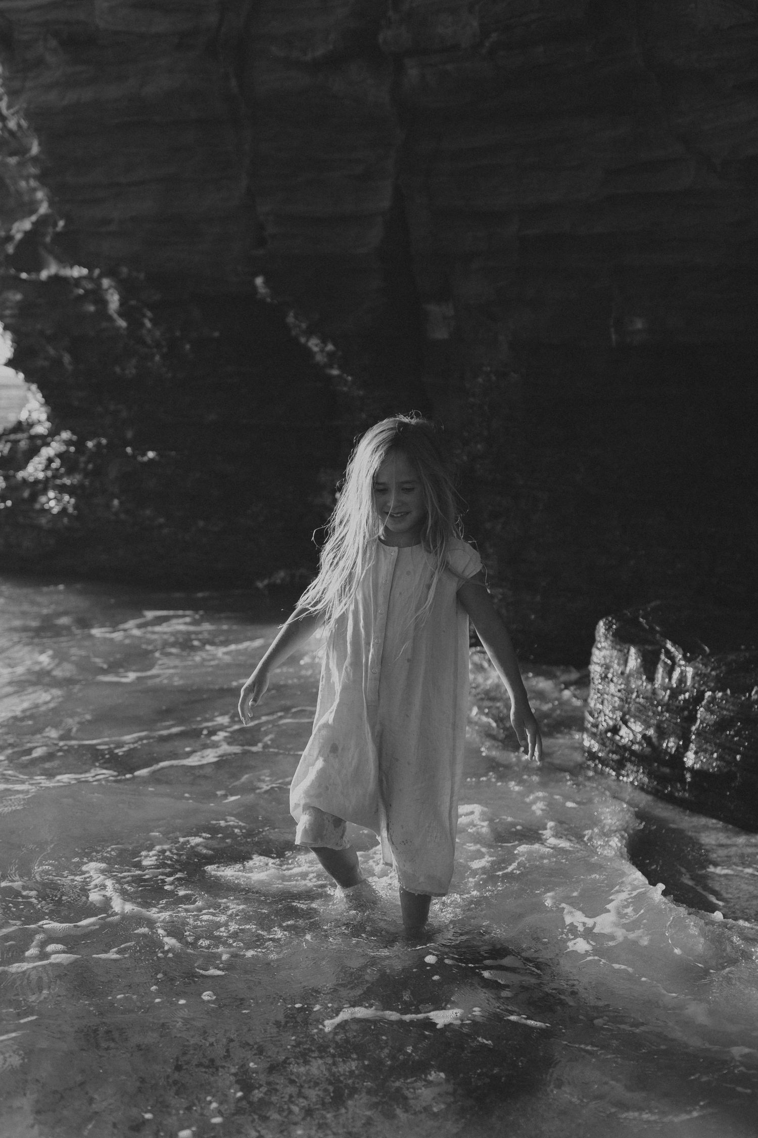 Person in white dress wading through shallow ocean water near rocky cliffs in black and white.