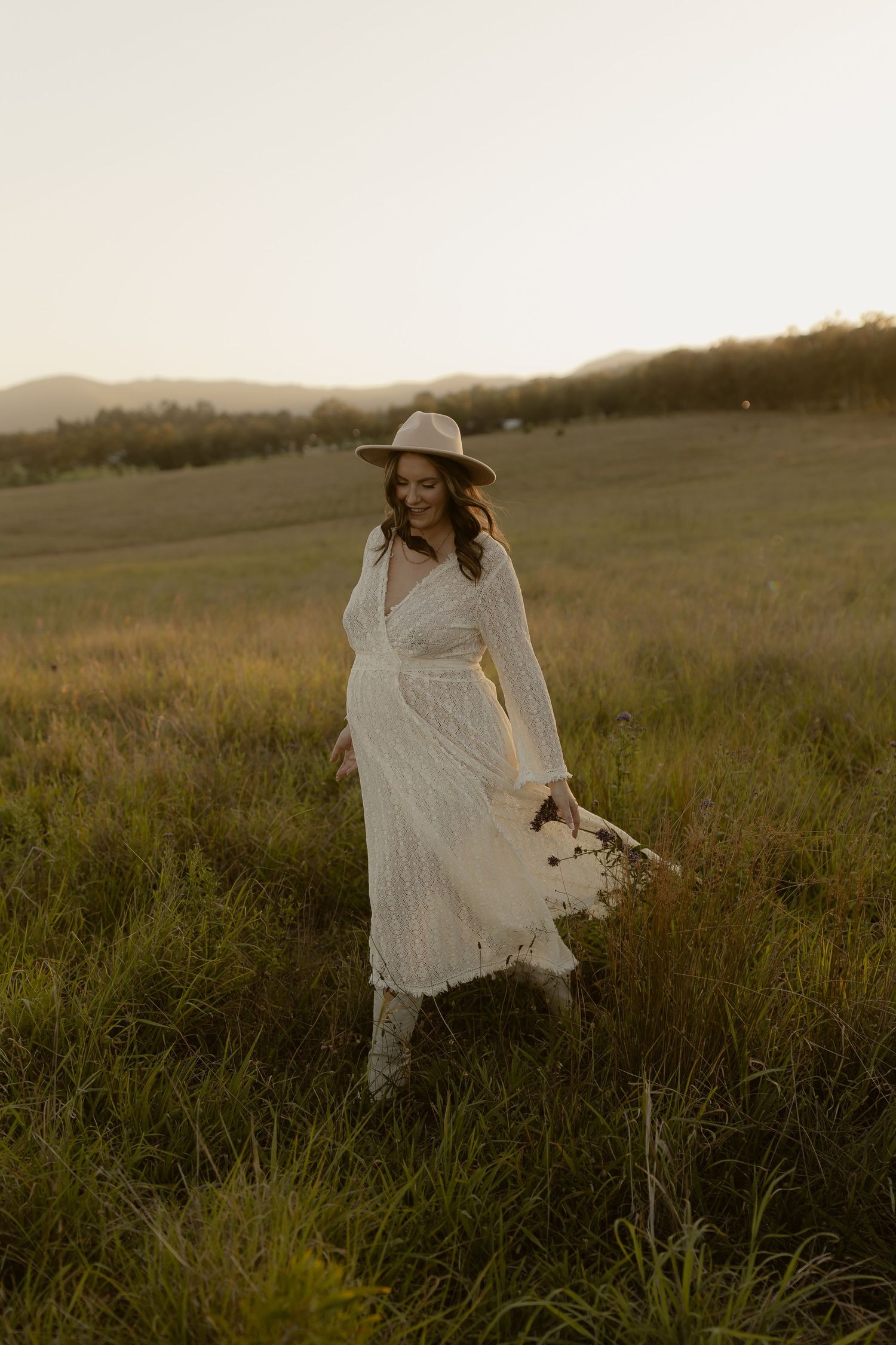 A person wearing a wide-brimmed hat and white dress moves gracefully through a sunlit field at dusk.