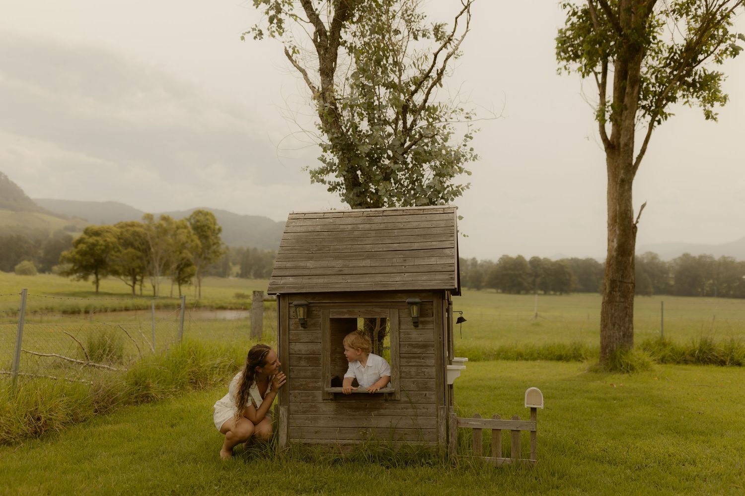 Two children play in a rustic wooden playhouse on a grassy field with trees and mountains in the background.