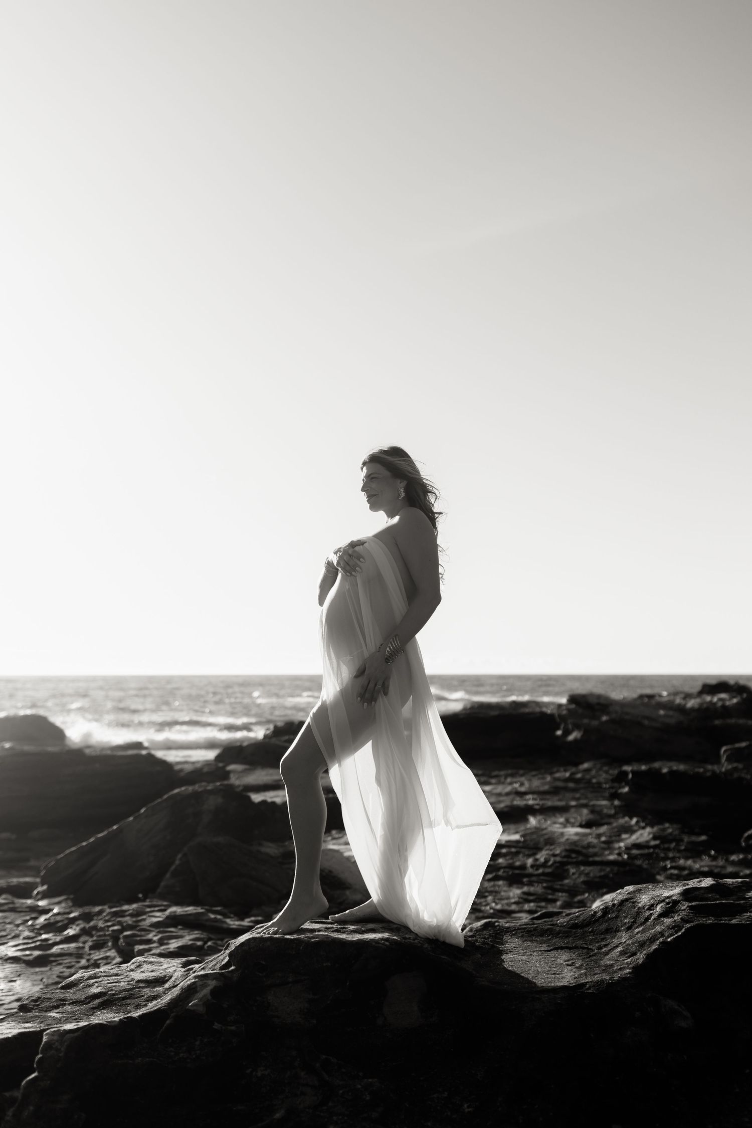 Black and white photo of a pregnant woman in sheer white fabric standing on coastal rocks with ocean waves behind her.
