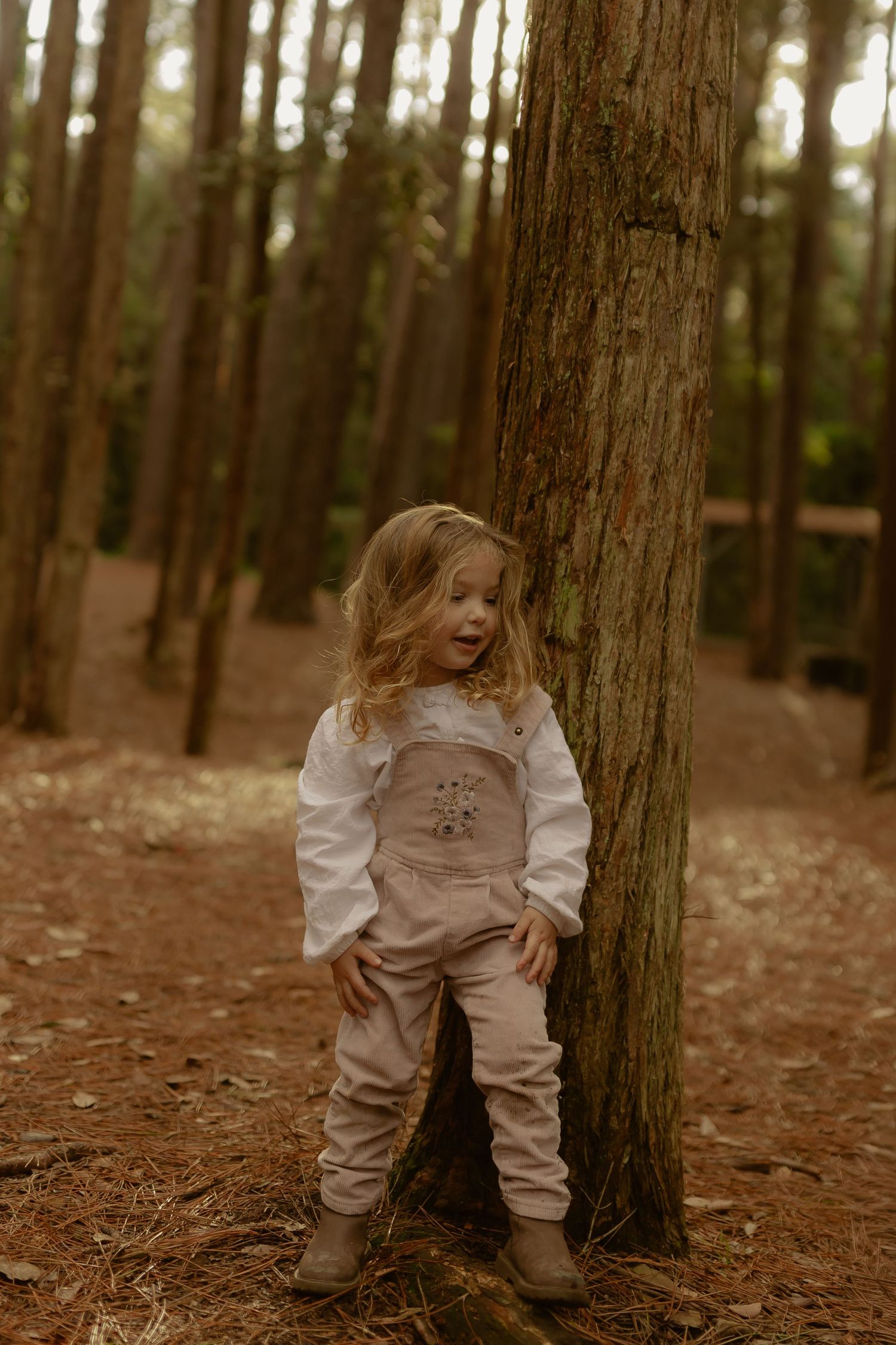 Curly-haired toddler girl in pink overalls leans against pine tree in autumn forest.