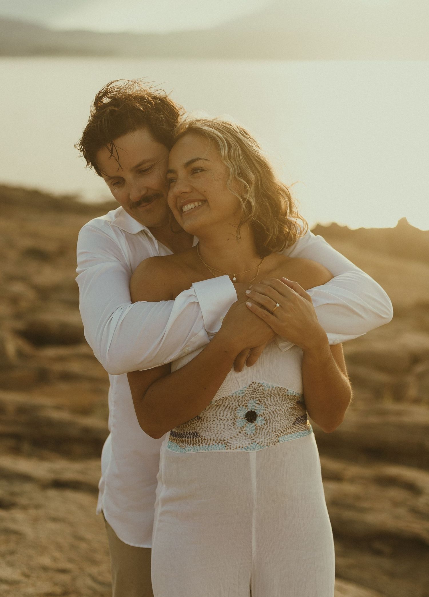 A romantic couple embraces on a coastal cliff during golden hour in matching white outfits.