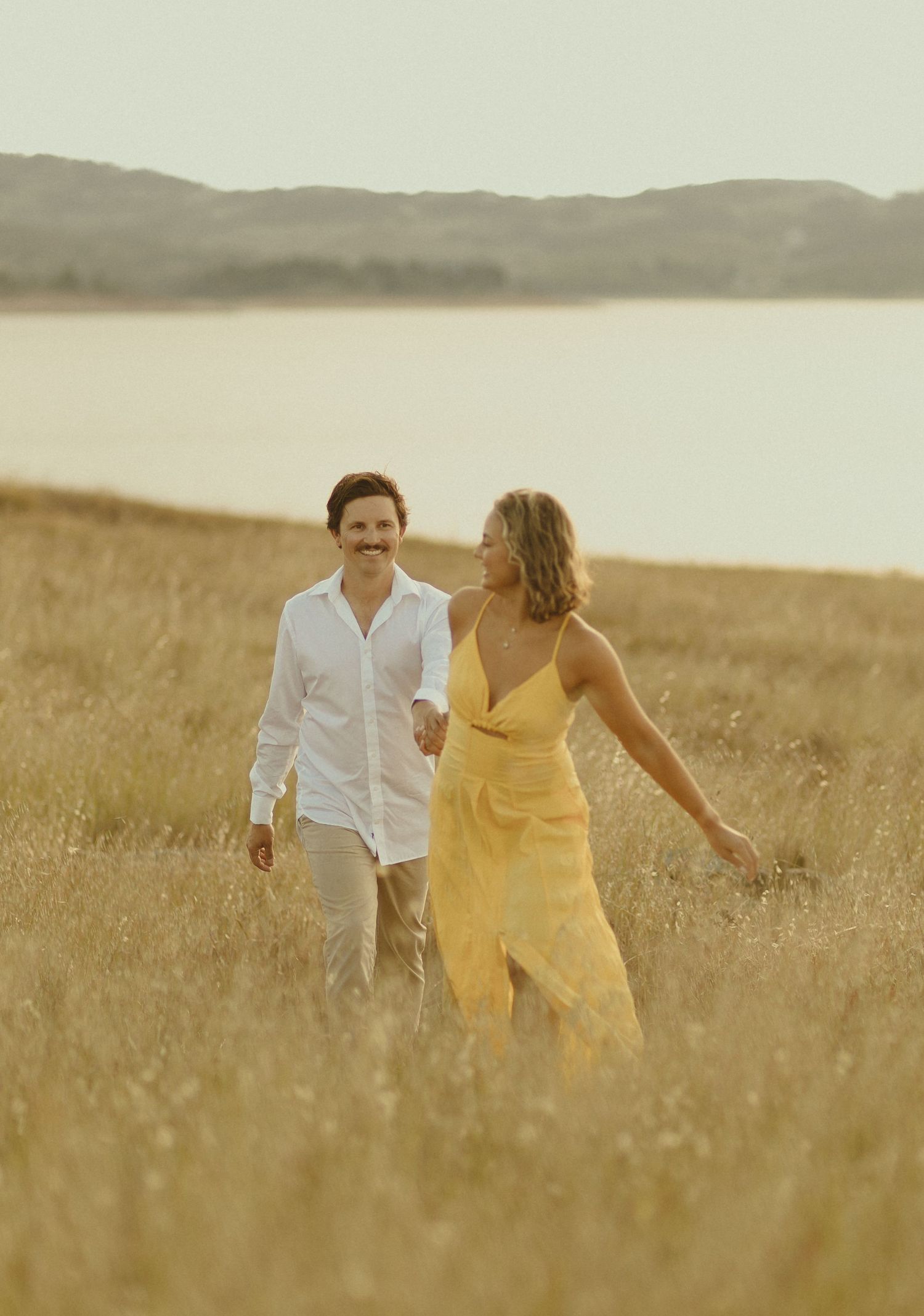 A romantic couple strolls through a sunlit meadow beside a lake, dressed in white and yellow summer attire.