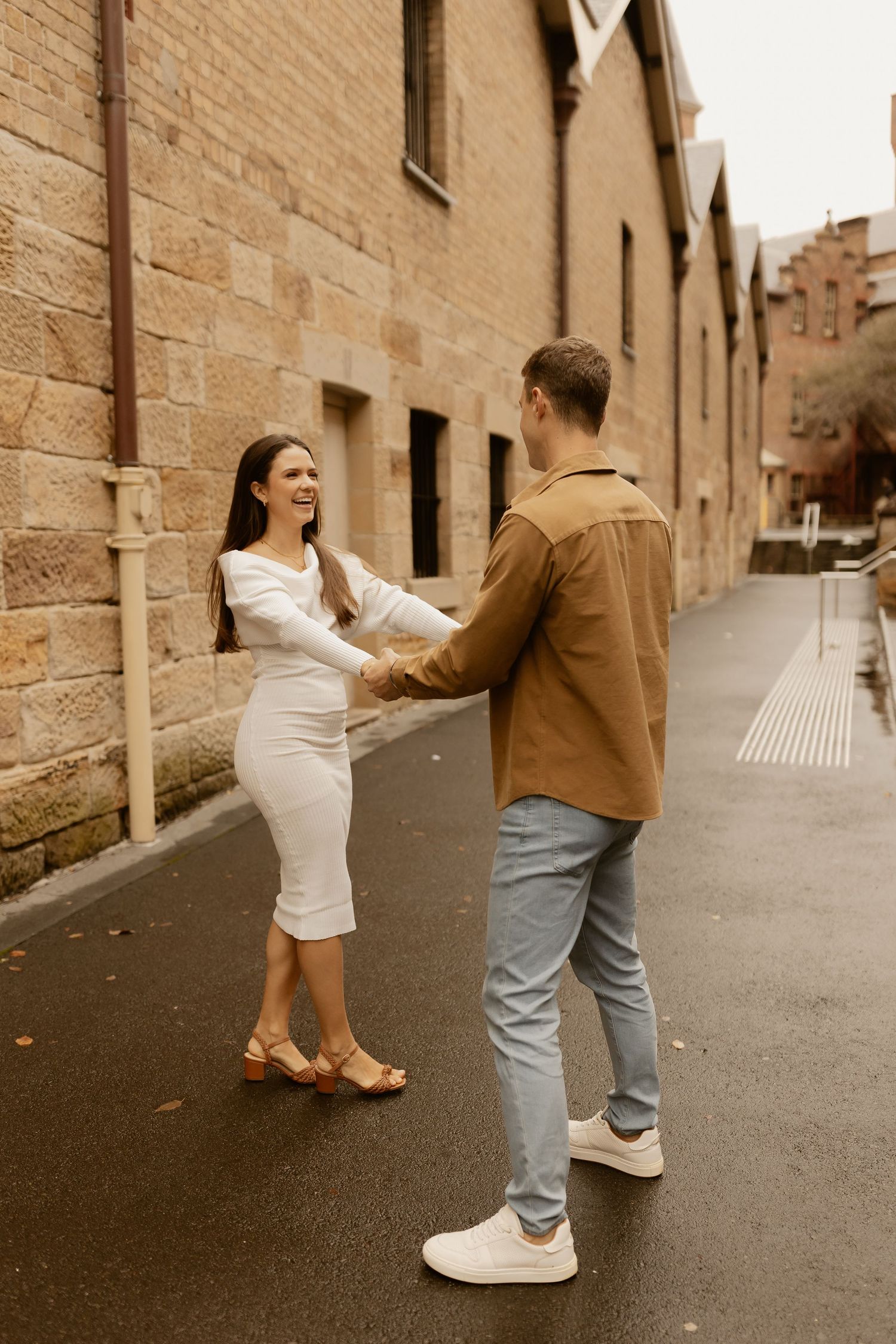 Couple in brown and white outfits dancing together on a cobblestone street beside brick buildings.
