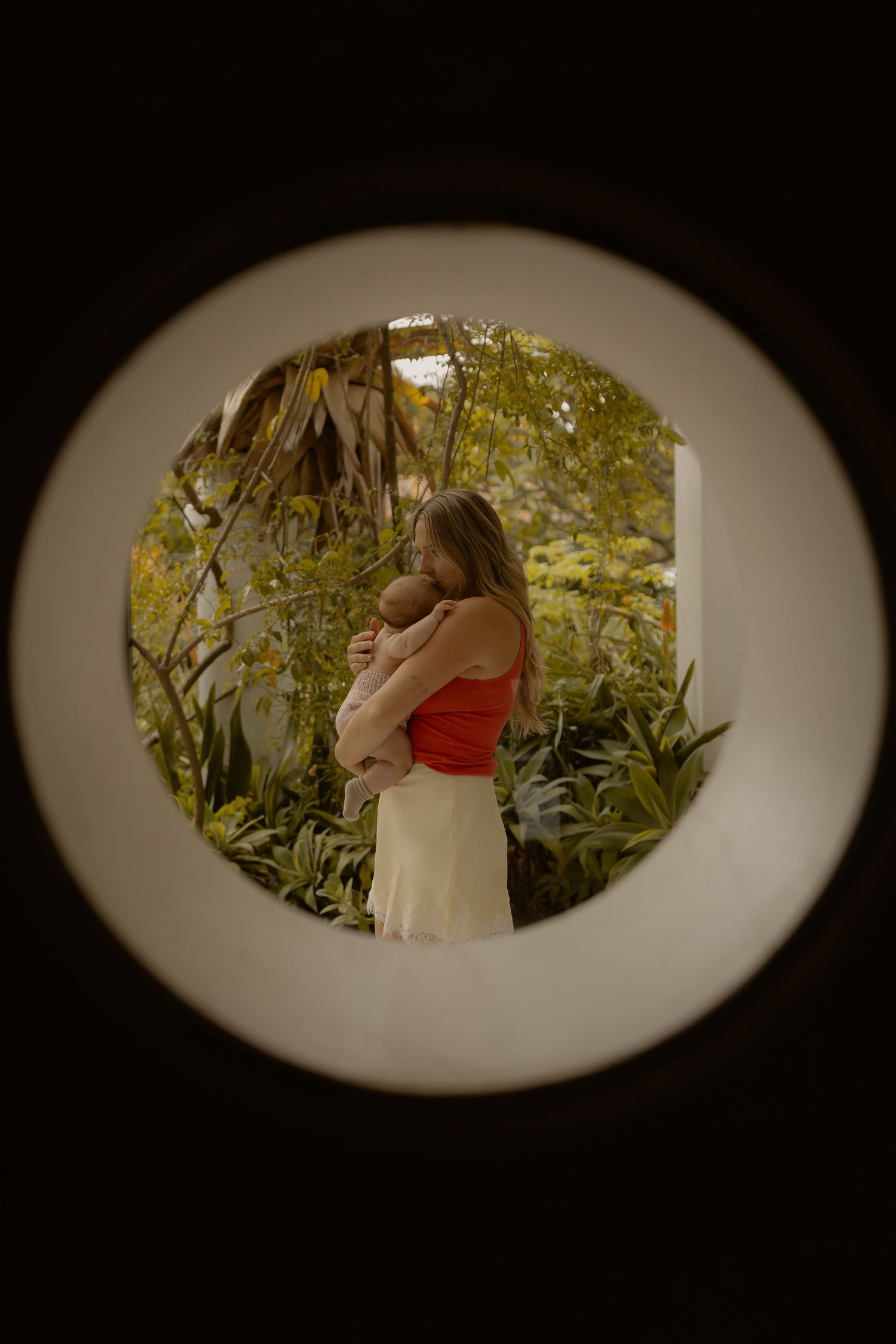 Woman in red top holding baby viewed through circular window surrounded by lush tropical garden greenery.