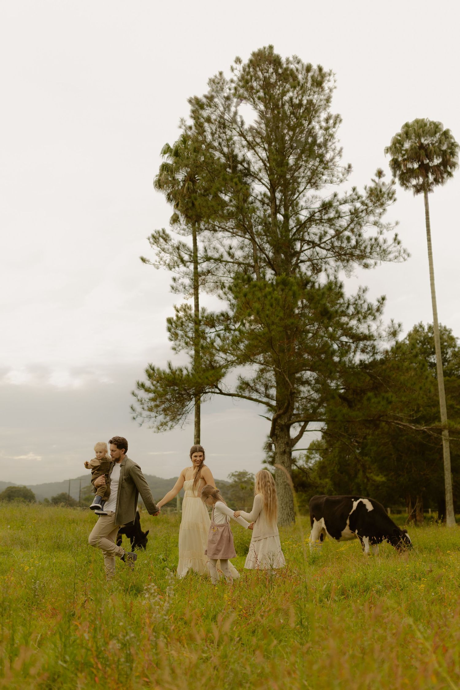 Family in white dresses and neutral tones walking through a green meadow with trees and a cow in the background.