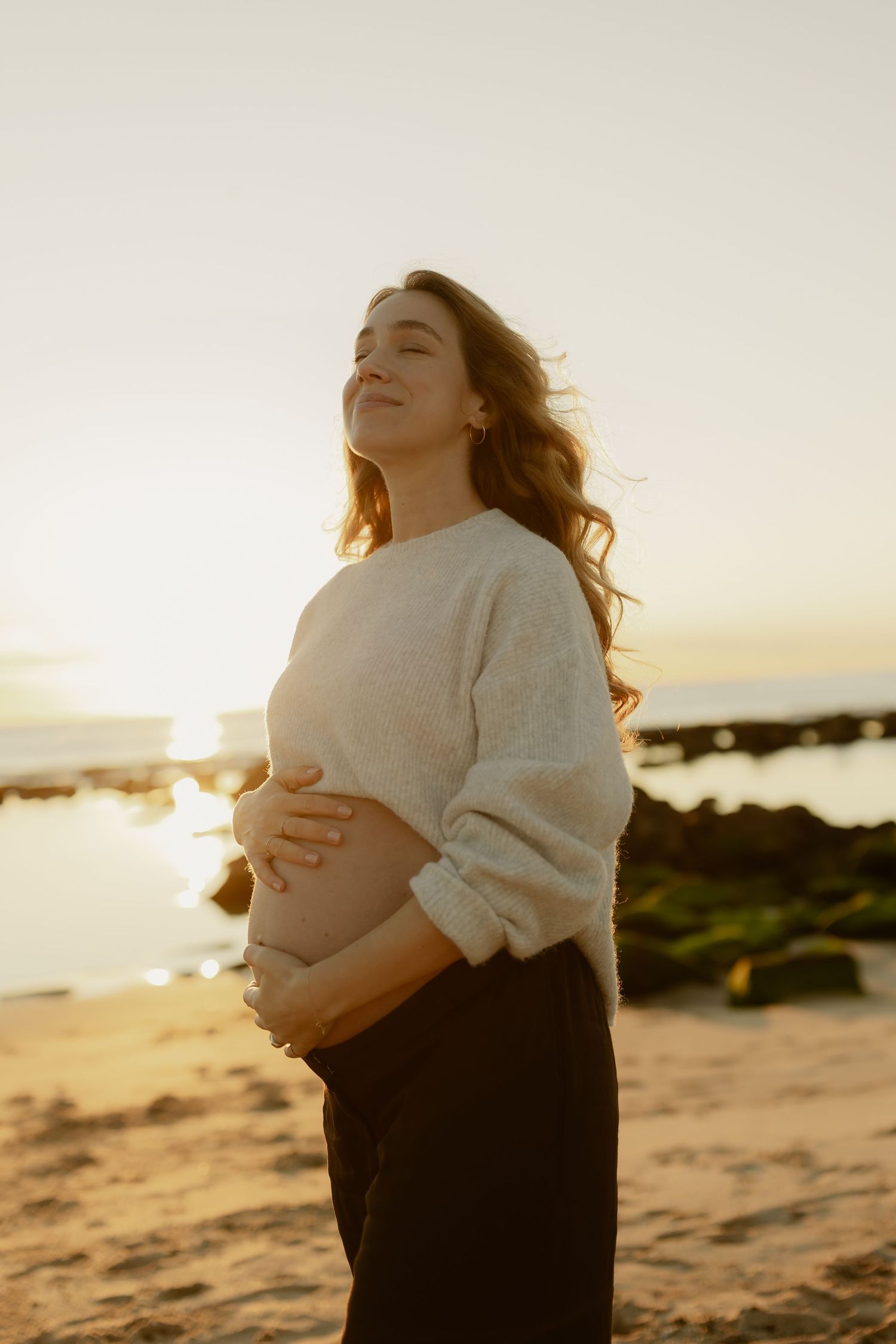 Pregnant woman in cream sweater cradling her baby bump on a sandy beach at golden sunset.