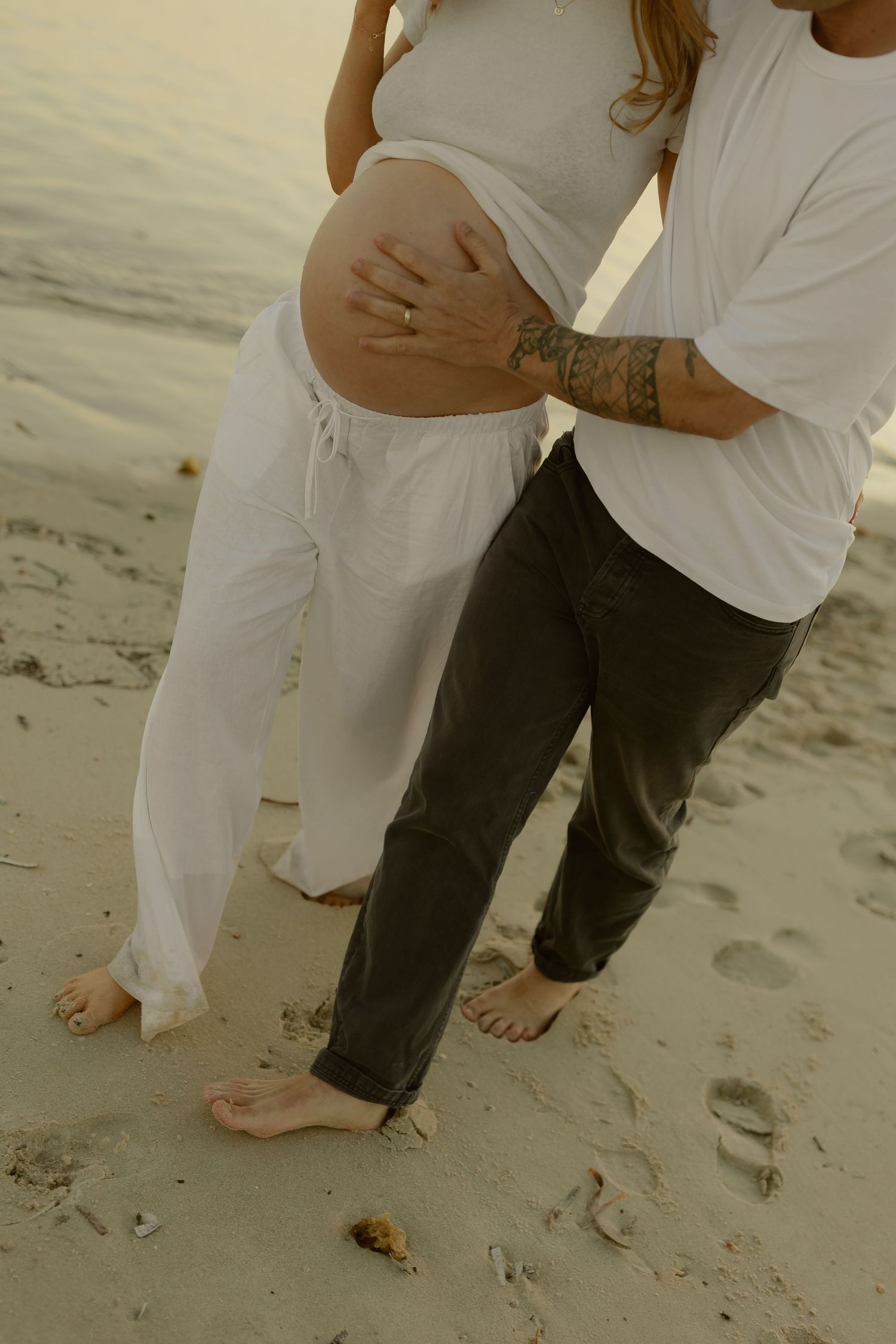 Couple on sandy beach, tattooed partner embracing pregnant womans bare bump, both barefoot in white and black.