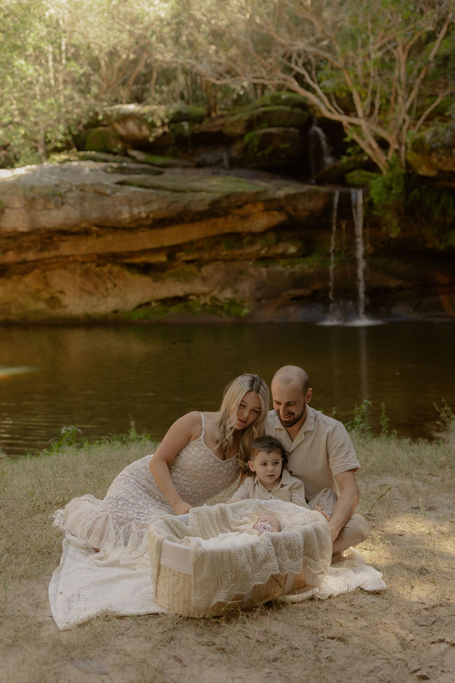 Family maternity photo session by a waterfall, parents and toddler gathered around a bassinet on a blanket.