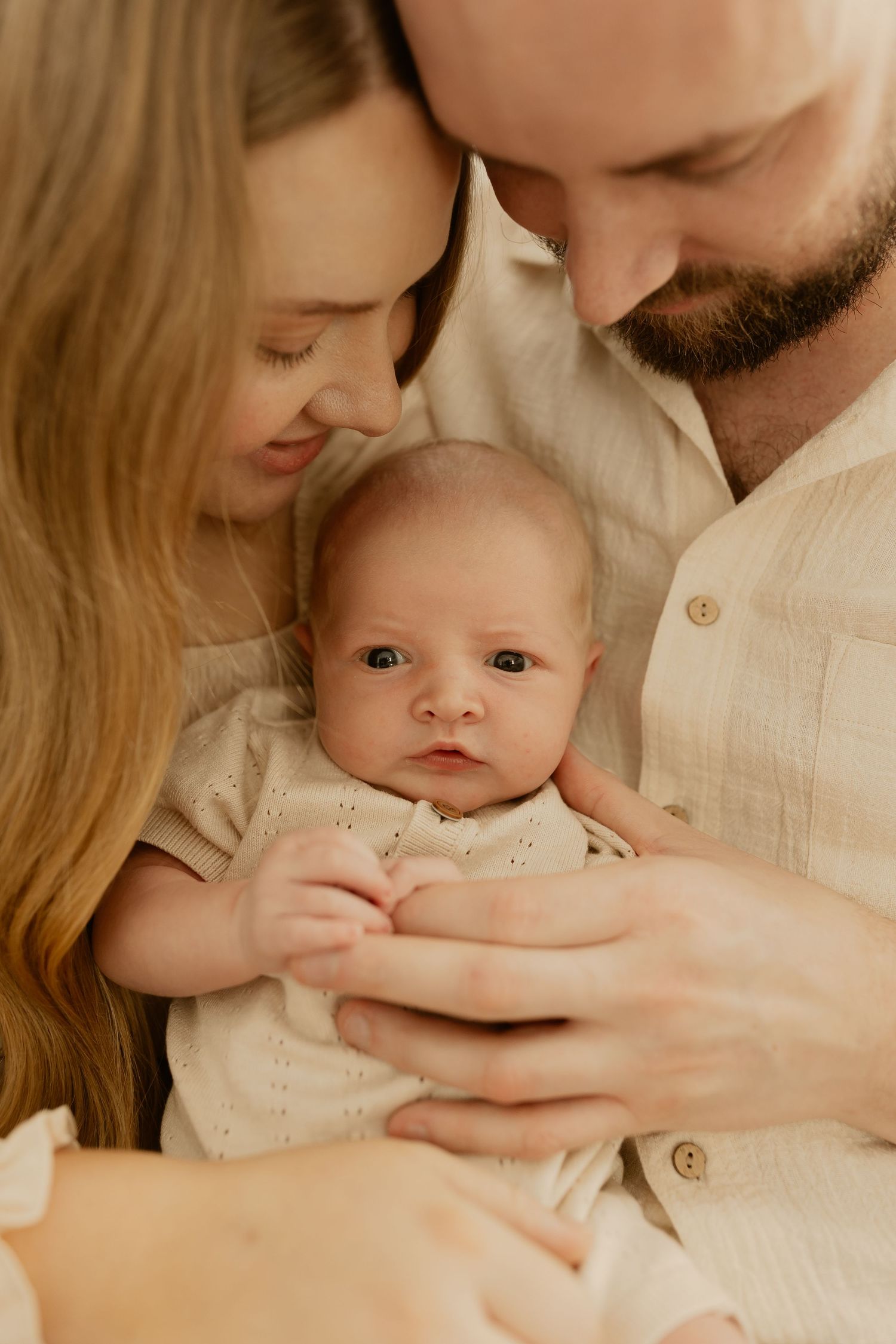 Parents in cream-colored clothing gaze lovingly at their alert baby in a warm, softly lit setting.