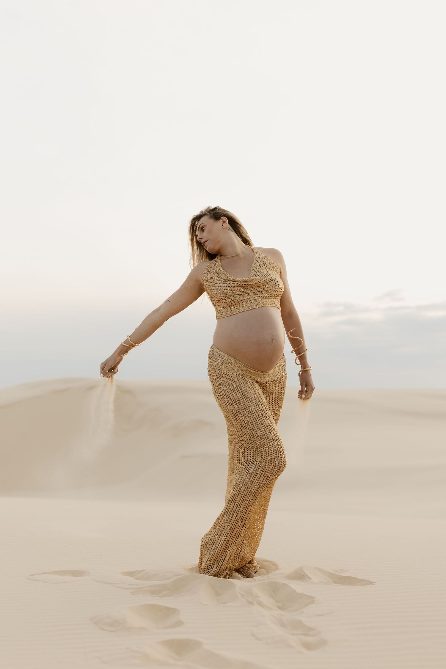 Figure wearing sheer gold dress dances expressively among desert sand dunes at dusk.