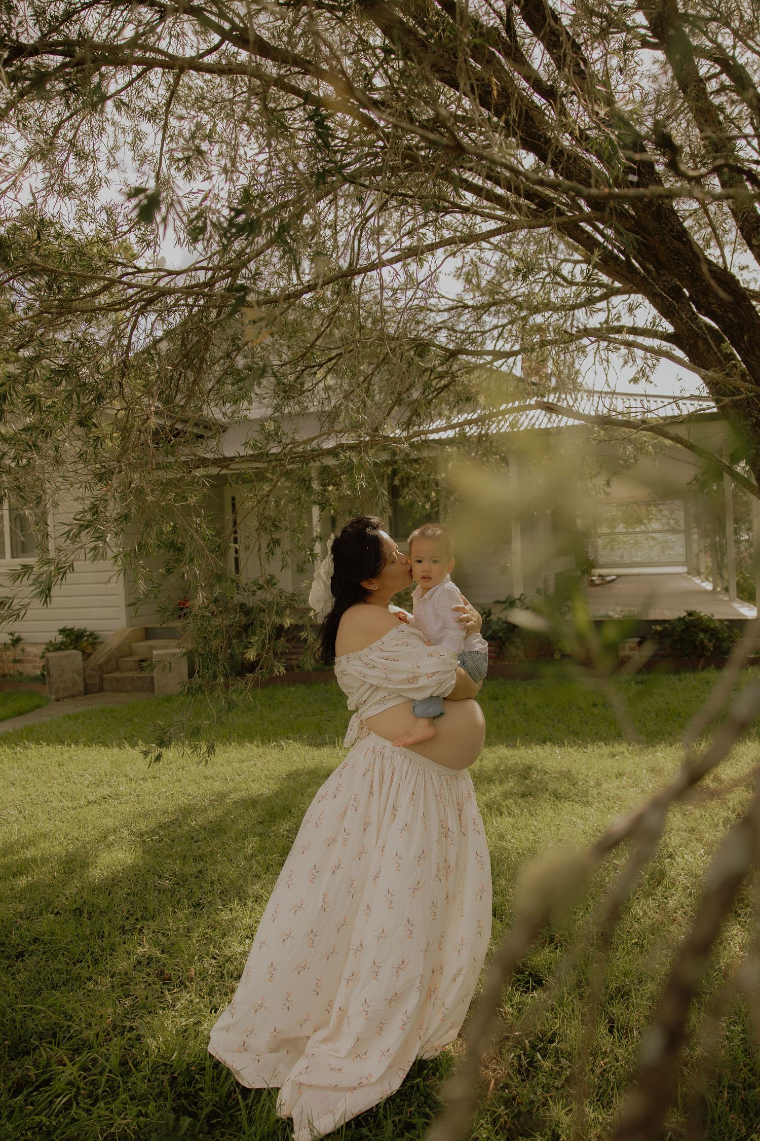 Pregnant woman in floral dress holding a baby under weeping tree branches outdoors.