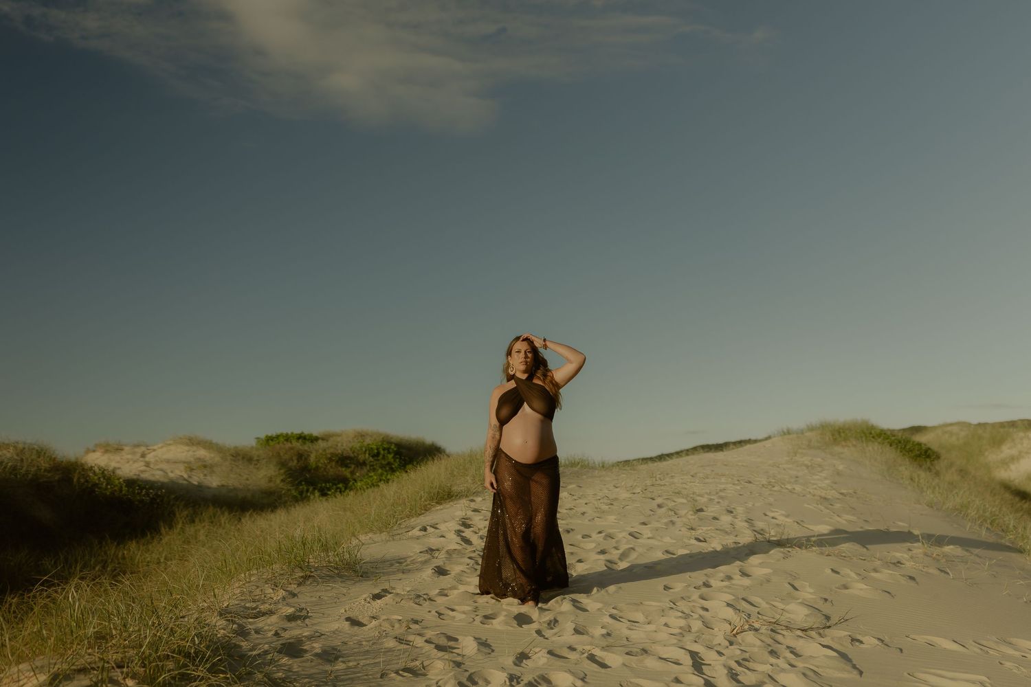 Pregnant woman in brown dress posing on sandy beach dunes under blue sky at golden hour.