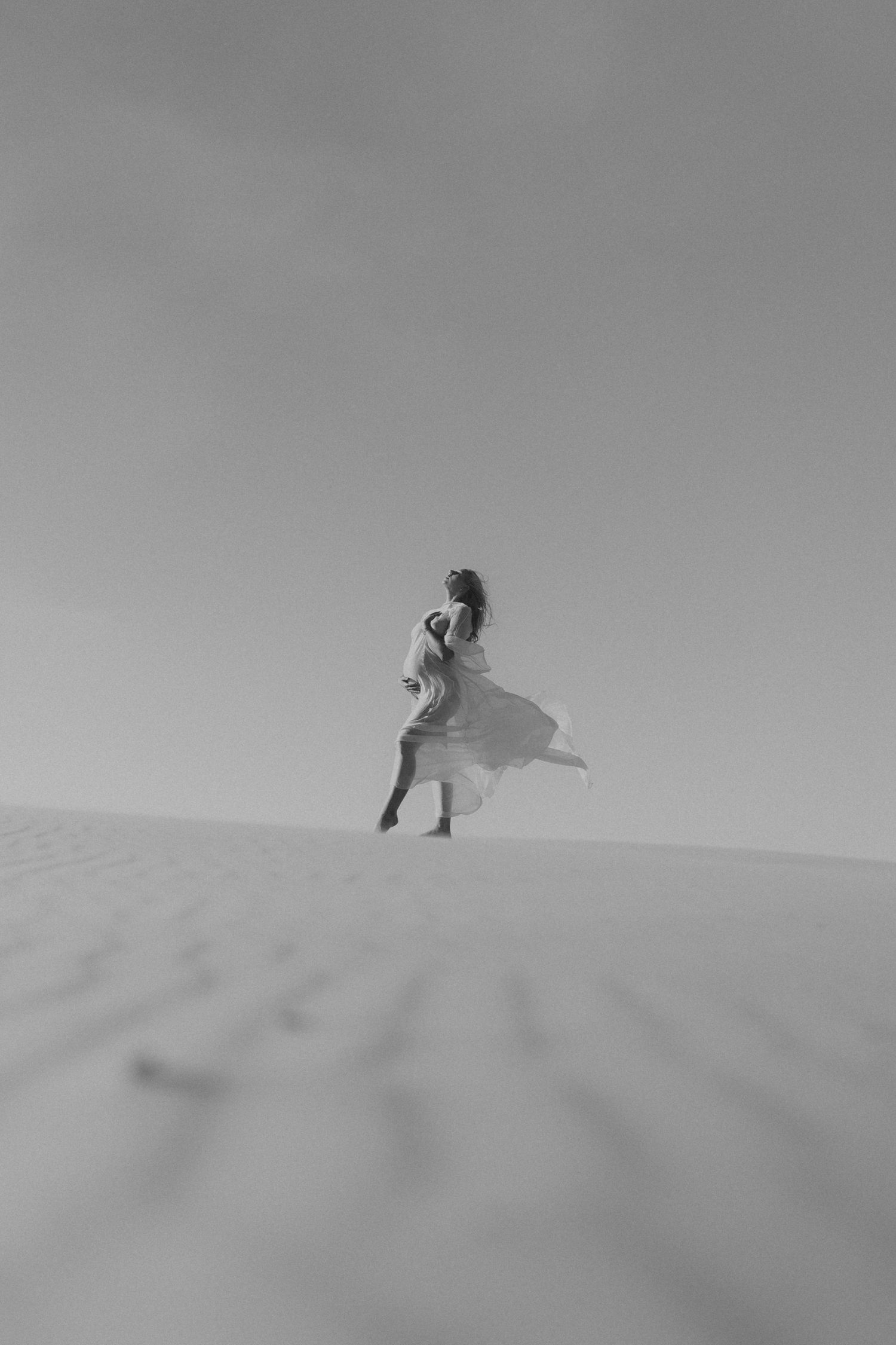 Black and white photo of a woman in a flowing white dress standing on a sand dune in a vast desert landscape.
