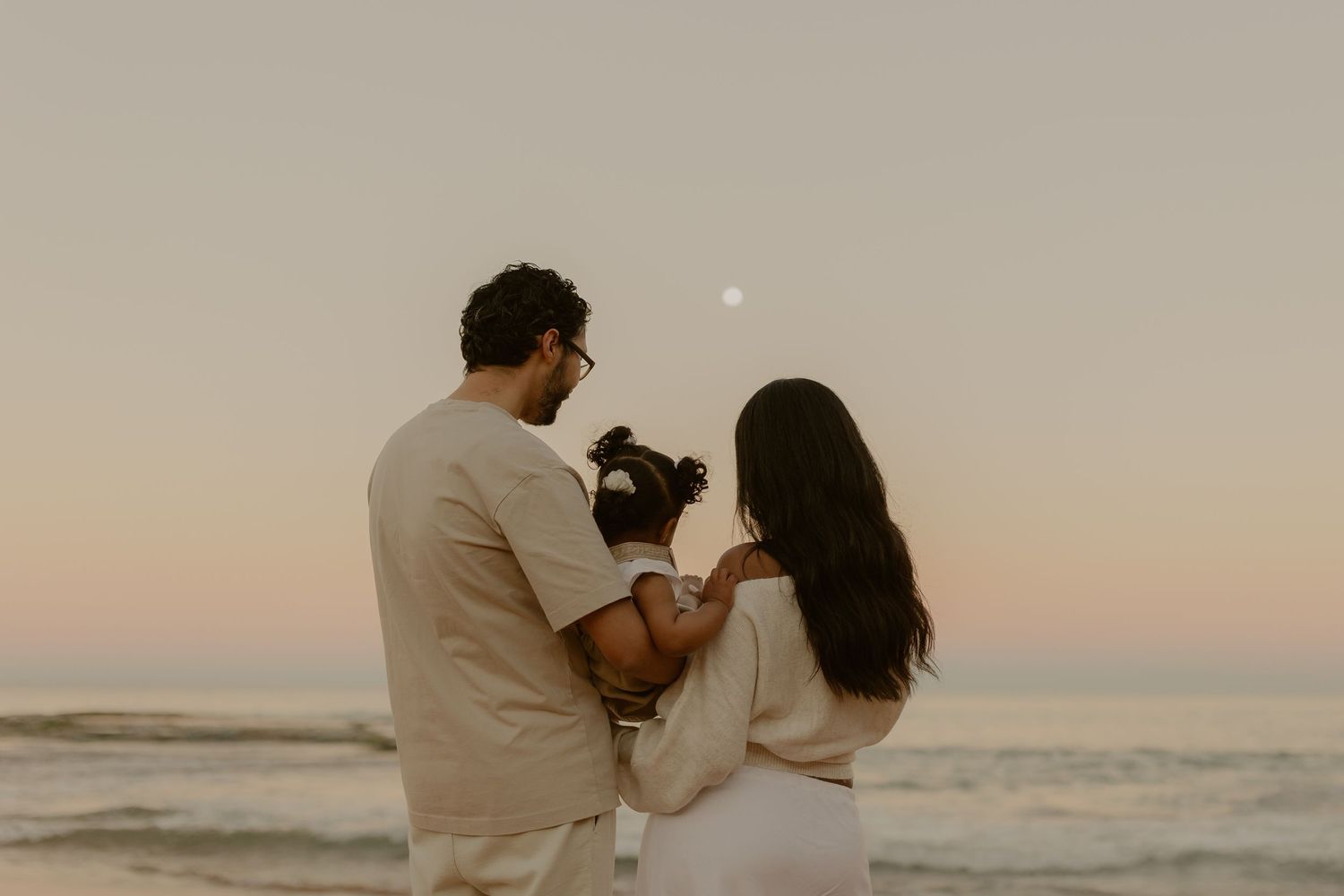 Couple standing on beach at sunset, photographing the moon rising over the ocean waves.