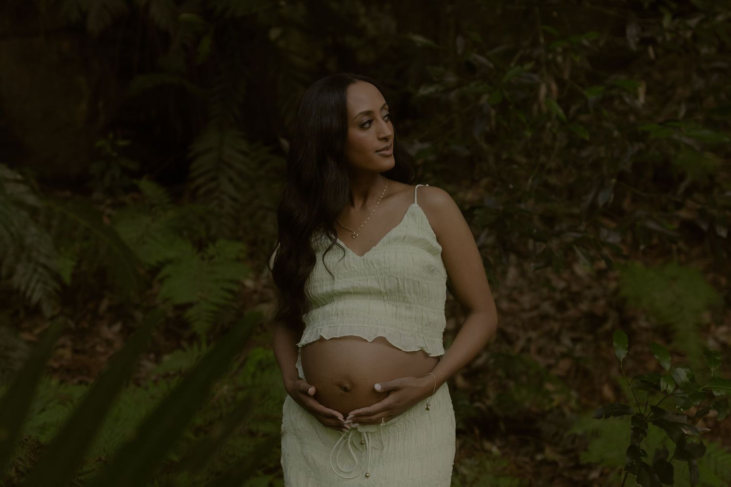 Pregnant woman in white dress cradling baby bump outdoors surrounded by lush green ferns in a forest setting.