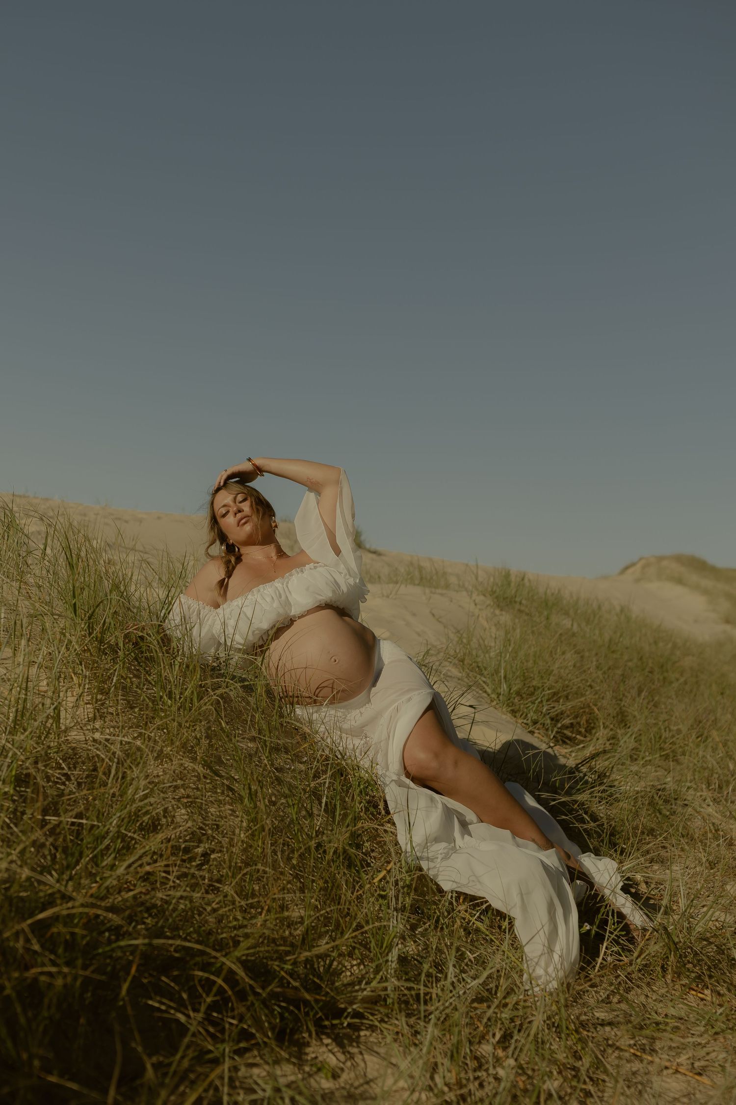 Pregnant woman in white dress reclines on sandy dunes with grass, posing gracefully under a clear blue sky.