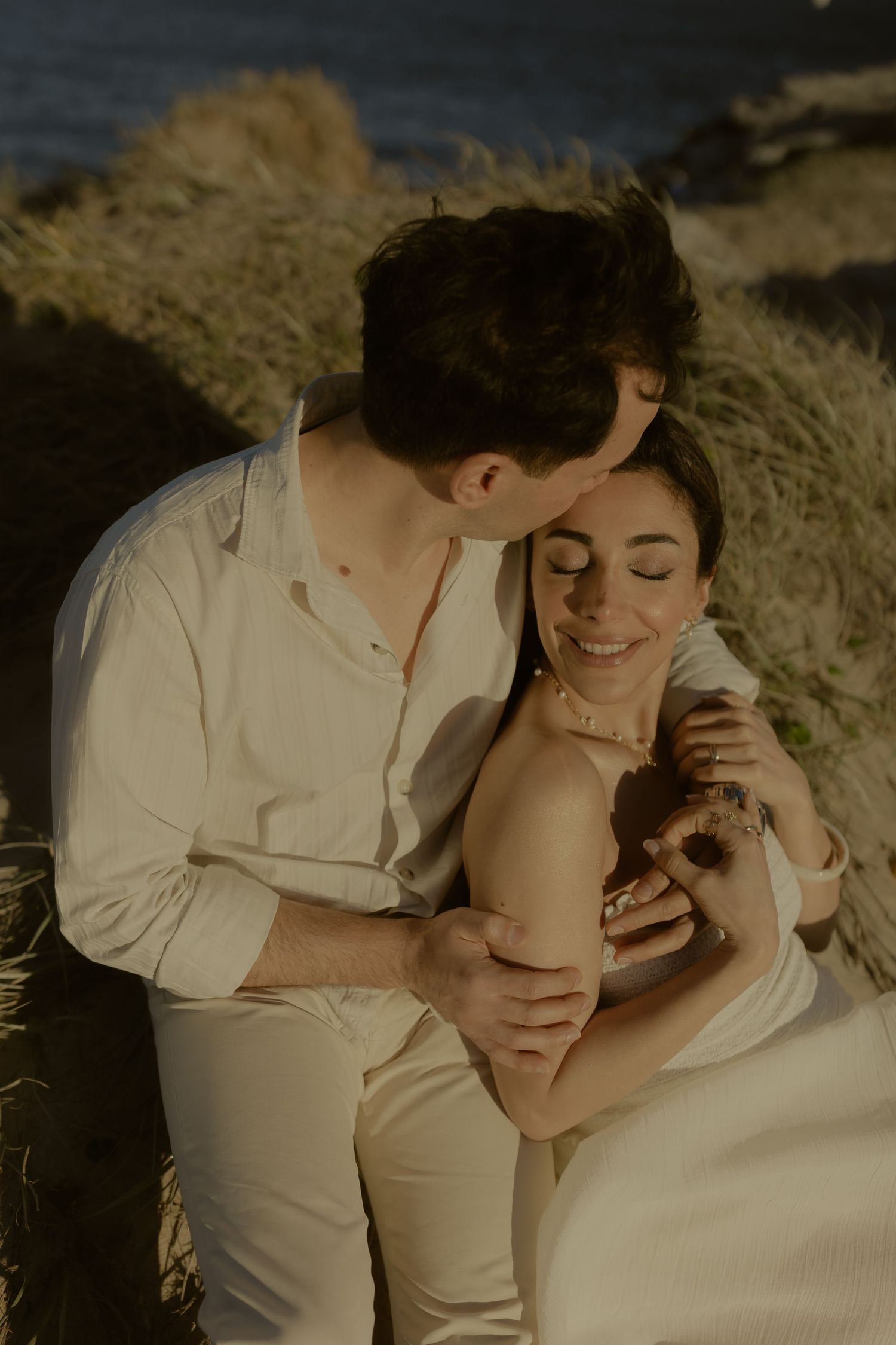 A couple sits together in golden grass near the ocean, sharing a tender embrace in matching white outfits.