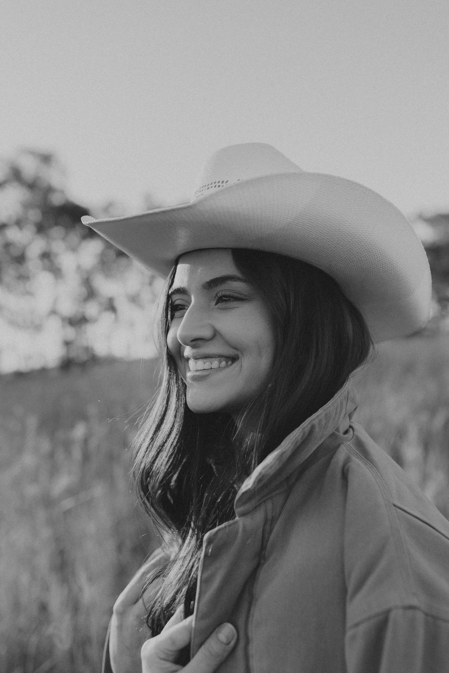 Smiling young woman wearing a cowboy hat in a black and white outdoor portrait photo.