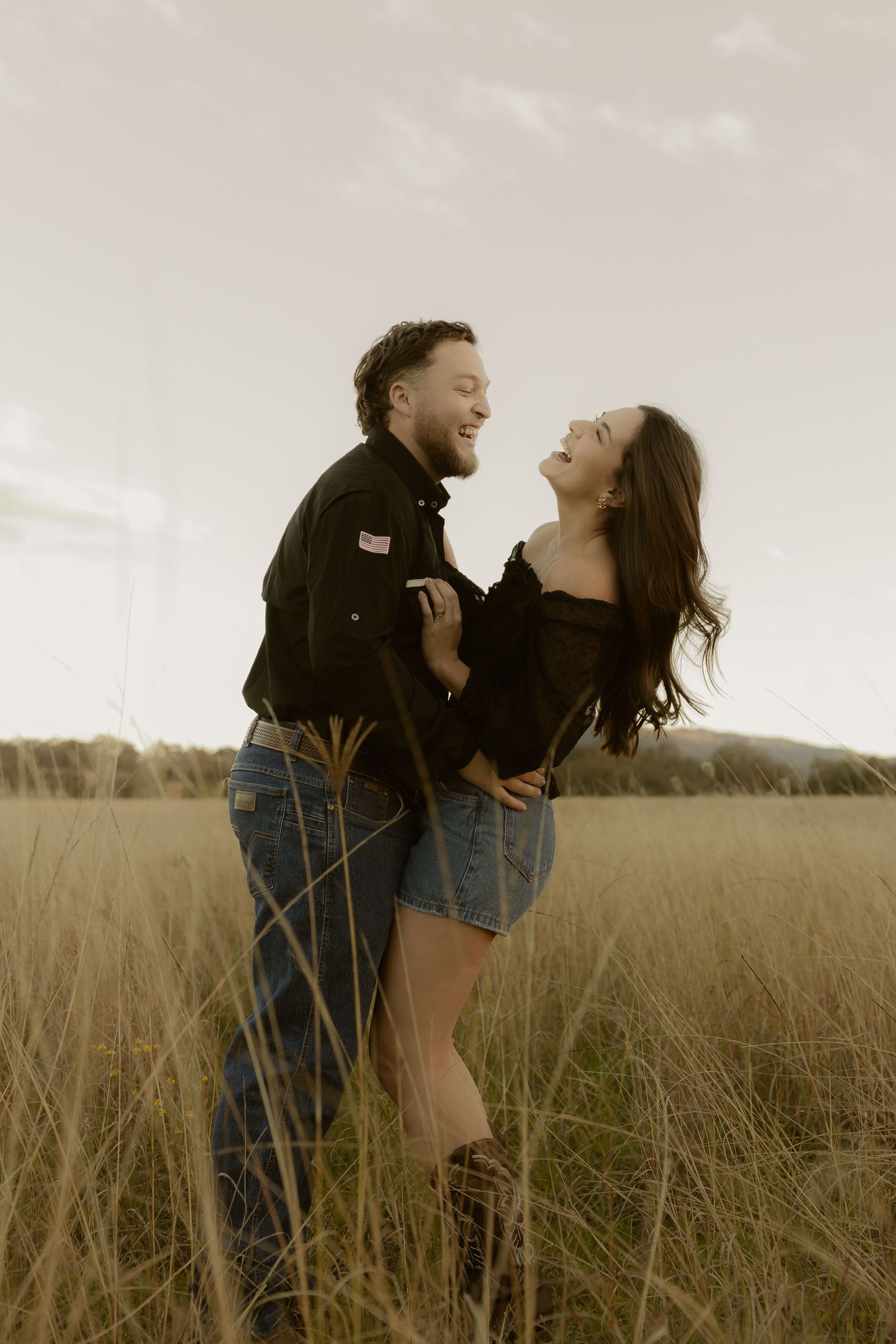 Happy couple laughing together while standing in tall golden grass field at sunset.