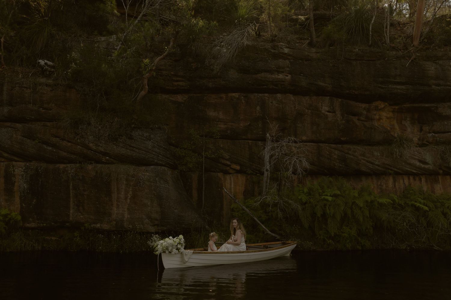A row of small white boats moored along a wooden dock in a tranquil wooded setting at dusk.