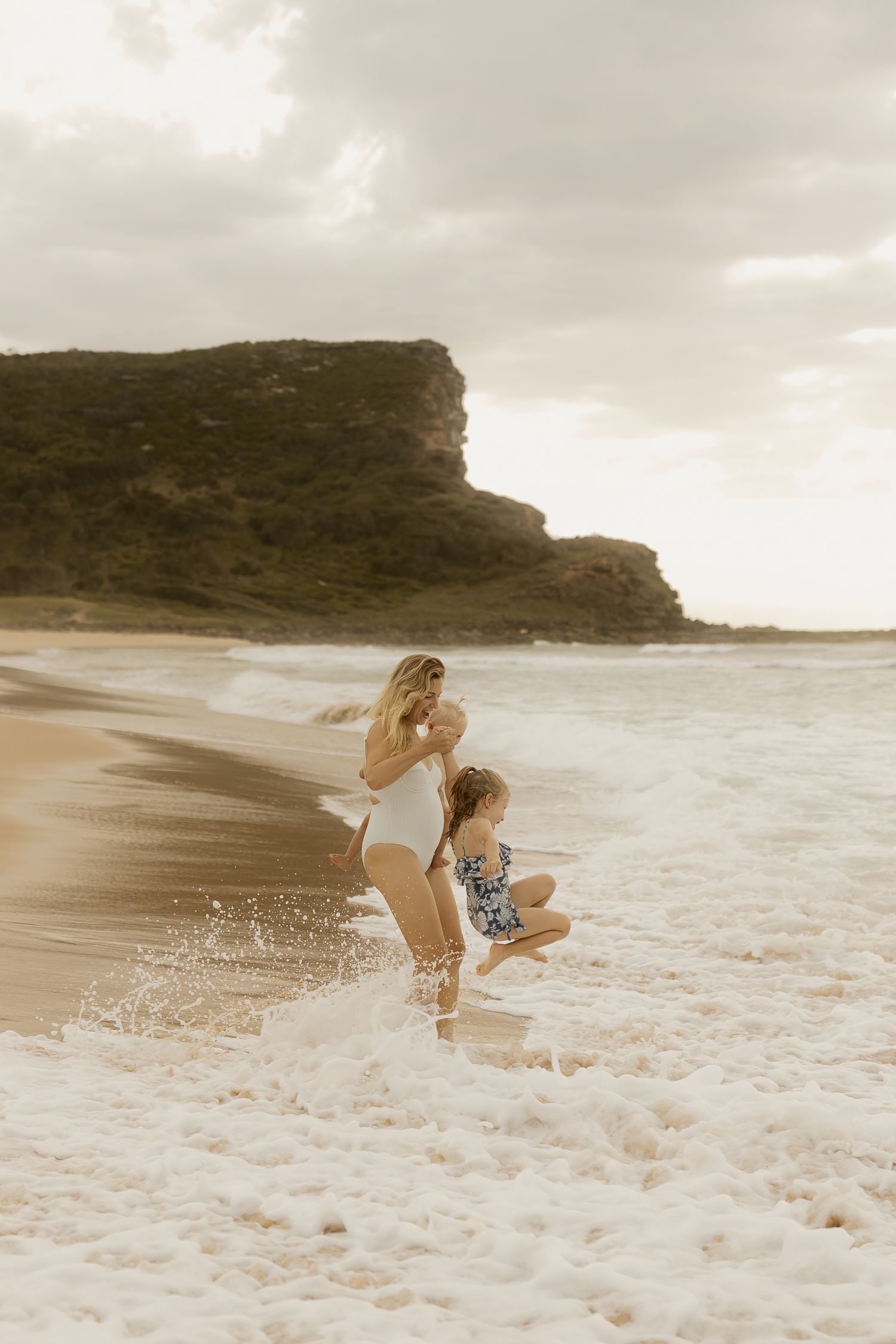 Two figures splash and play in the ocean waves against a dramatic coastal cliff backdrop at sunset.