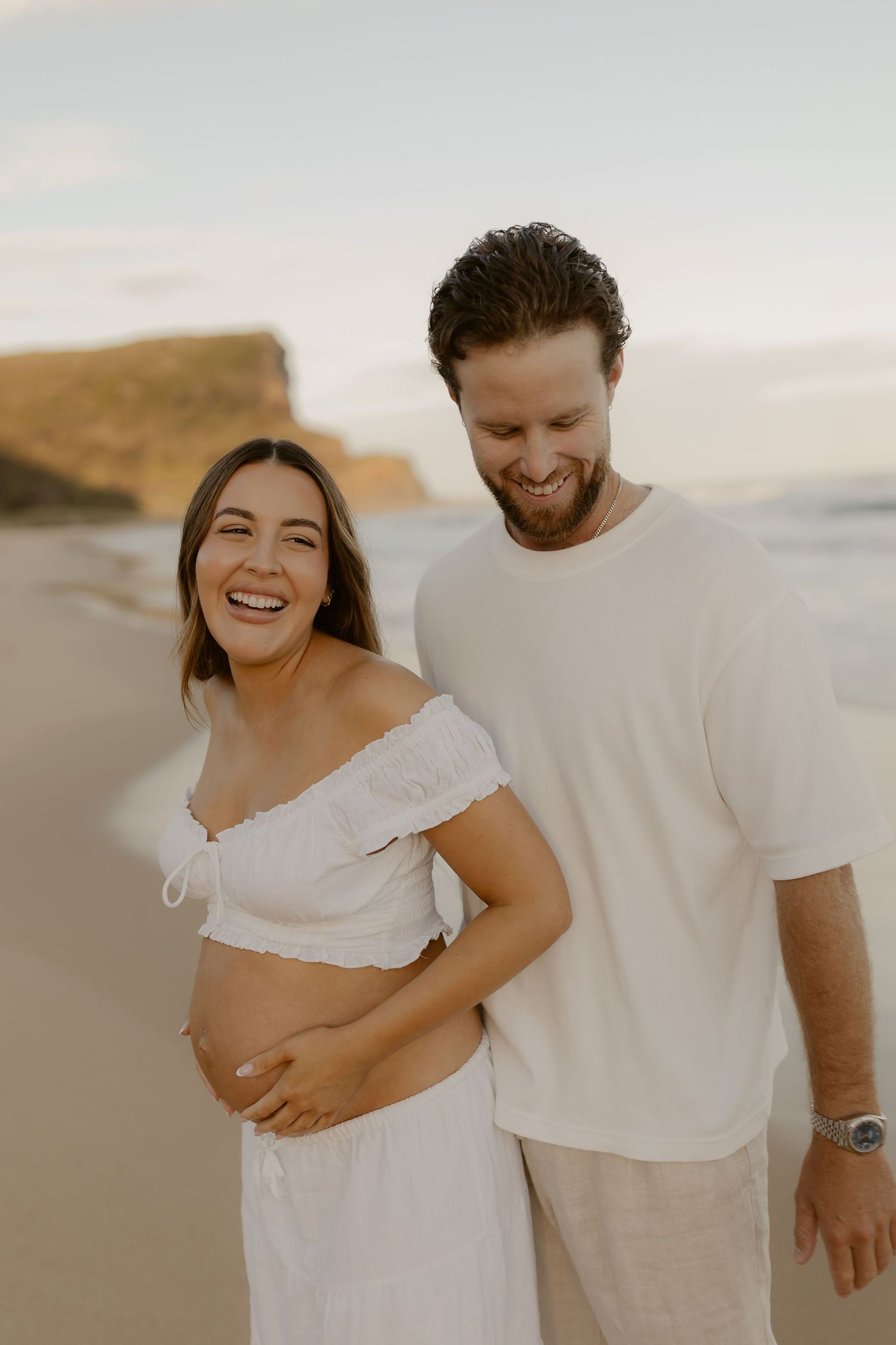 A couple shares joyful moments together on a beach at sunset wearing white casual outfits.