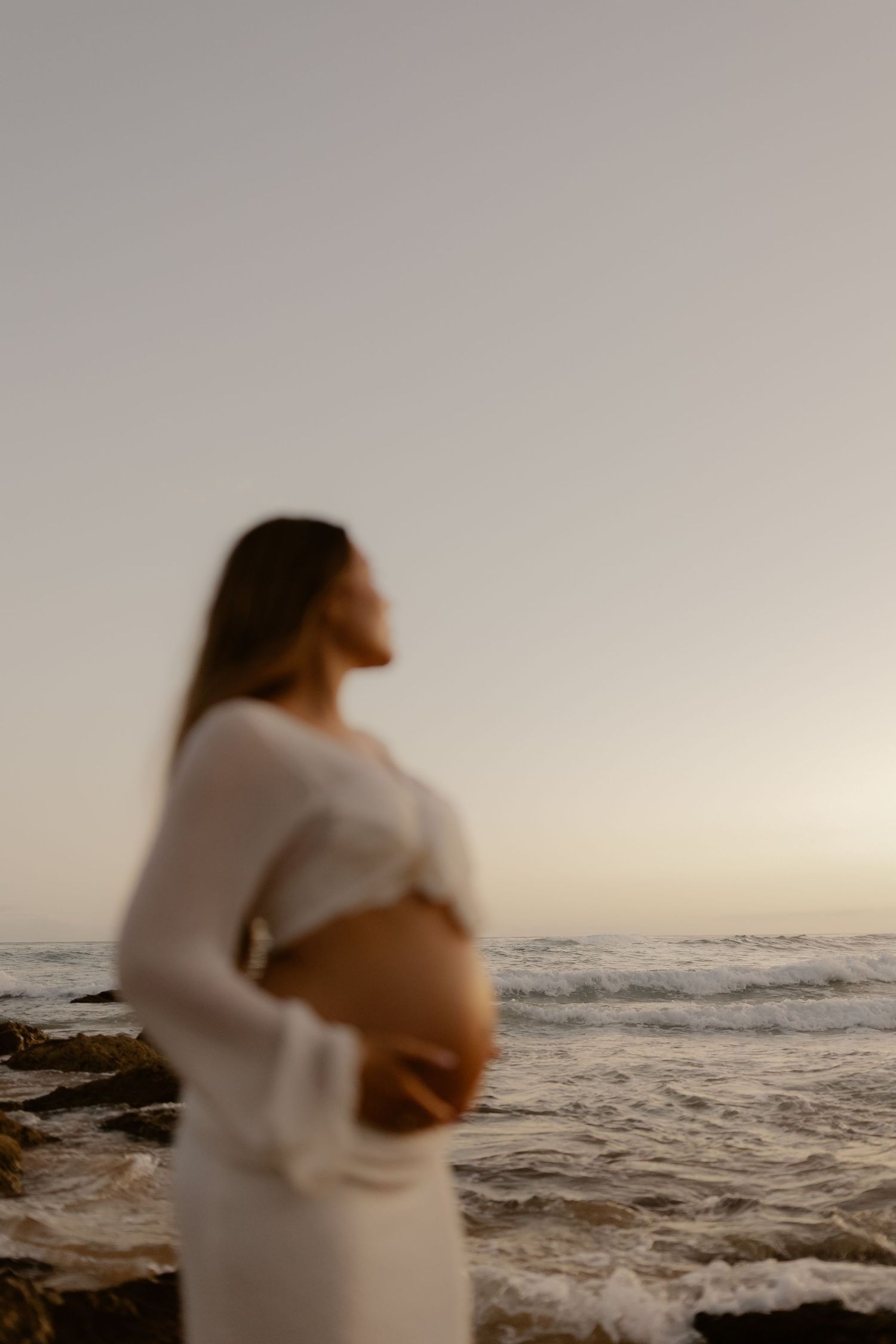 A pregnant woman in a white dress stands on a rocky beach at sunset, cradling her belly against an ocean backdrop.
