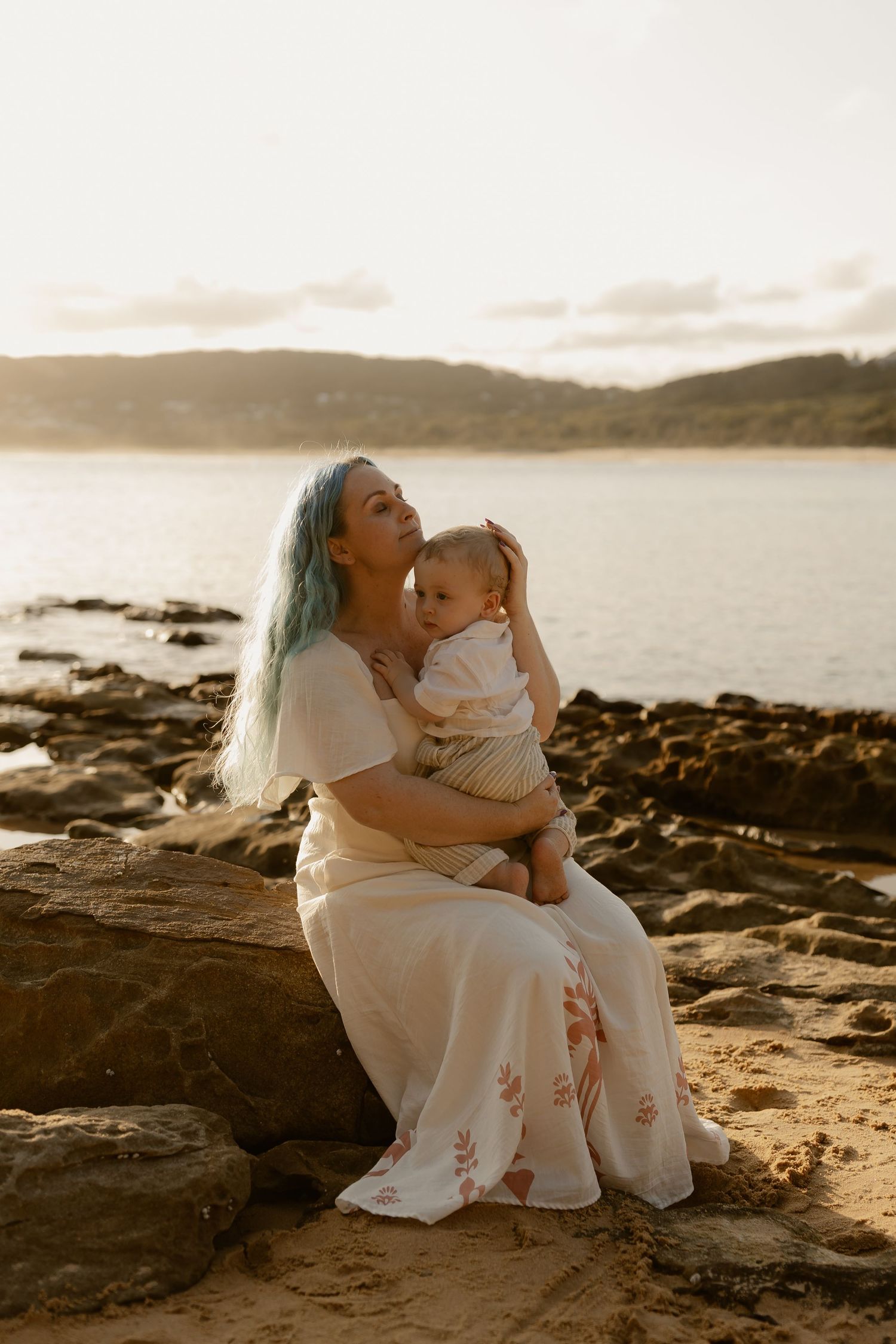 A tender moment captured at sunset on a rocky beach with ocean views in the background.