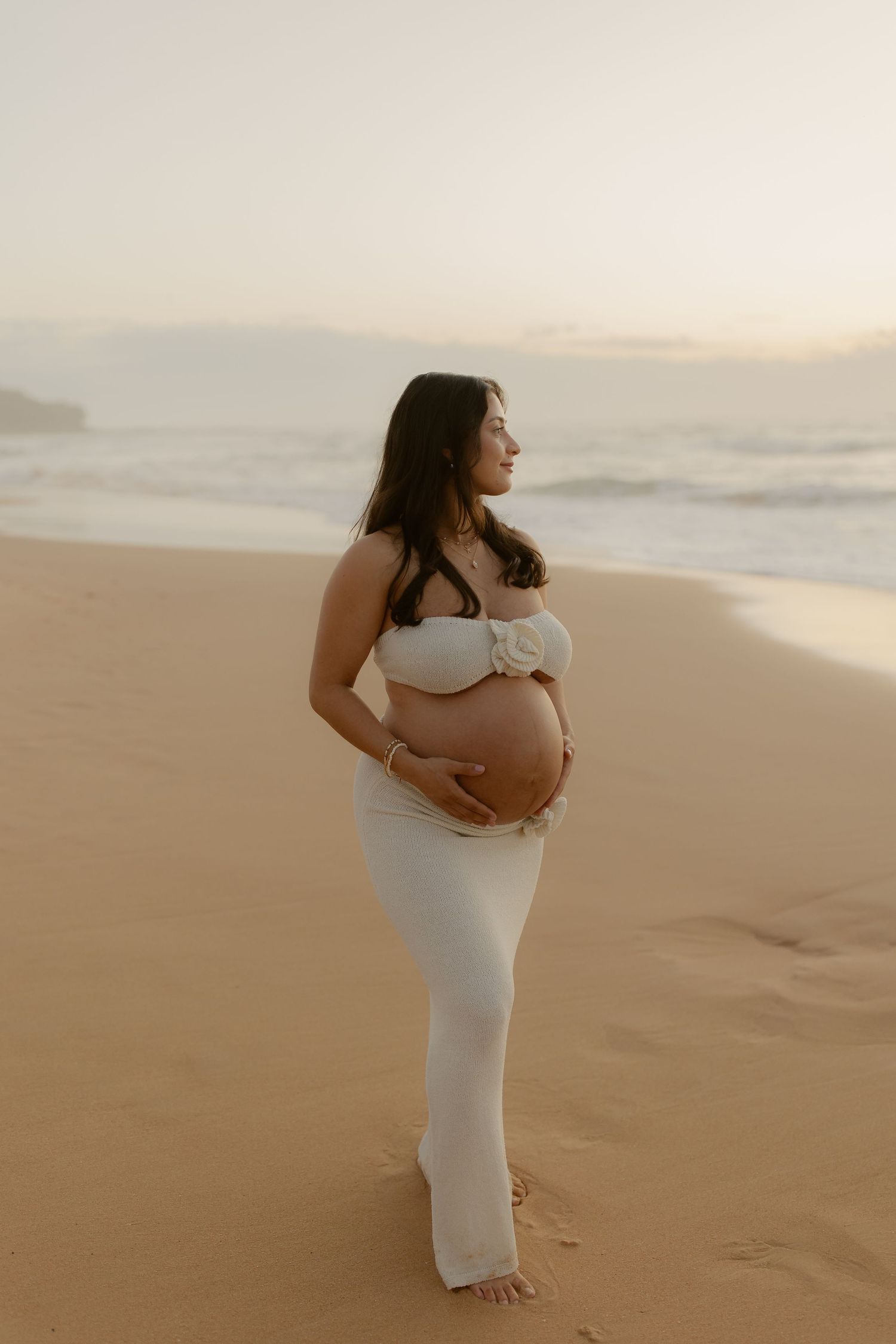 A woman in a flowing white dress stands on a sandy beach at sunset, creating a serene coastal silhouette.