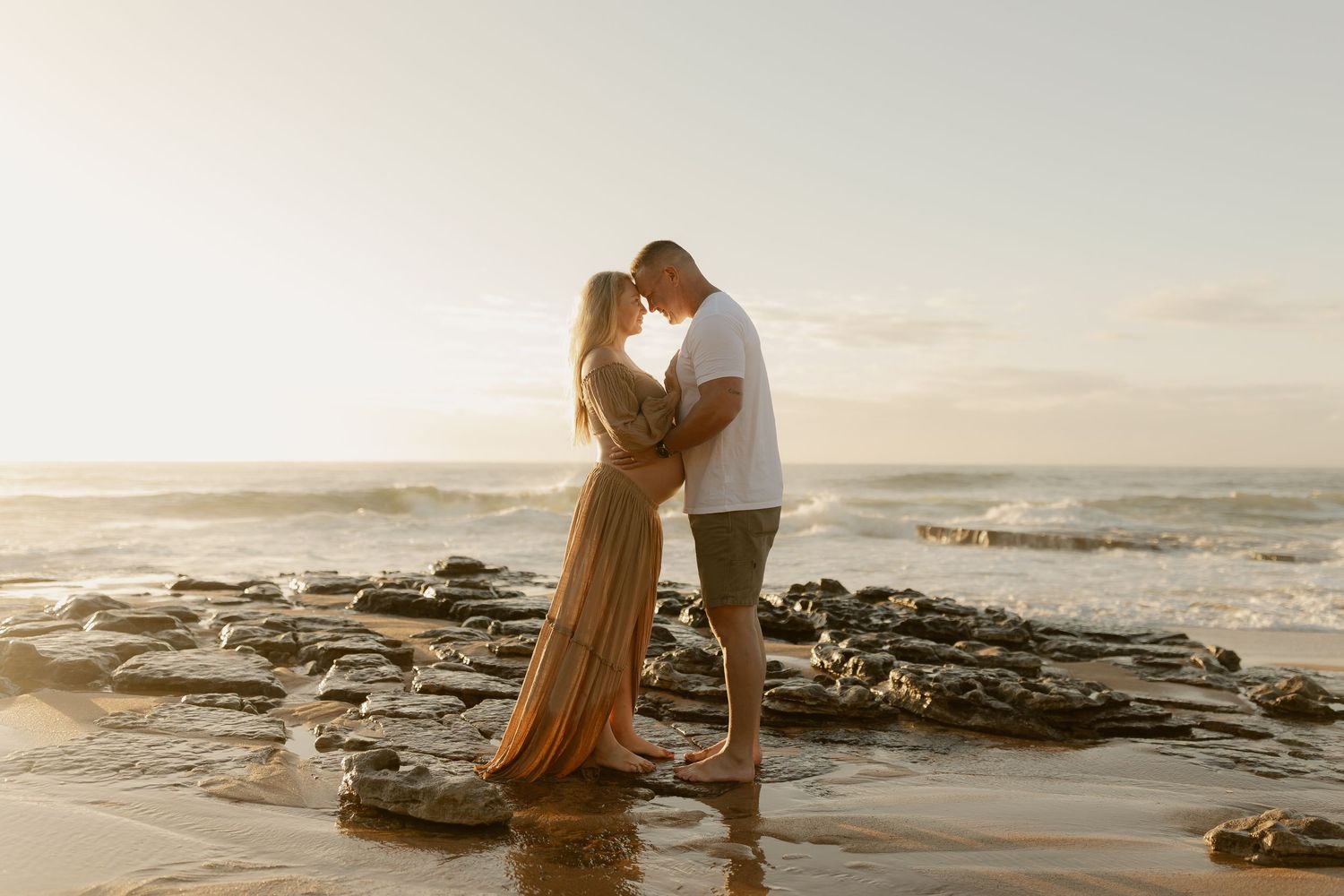Expecting couple sharing a romantic kiss on rocky shoreline with golden sunset light behind them.