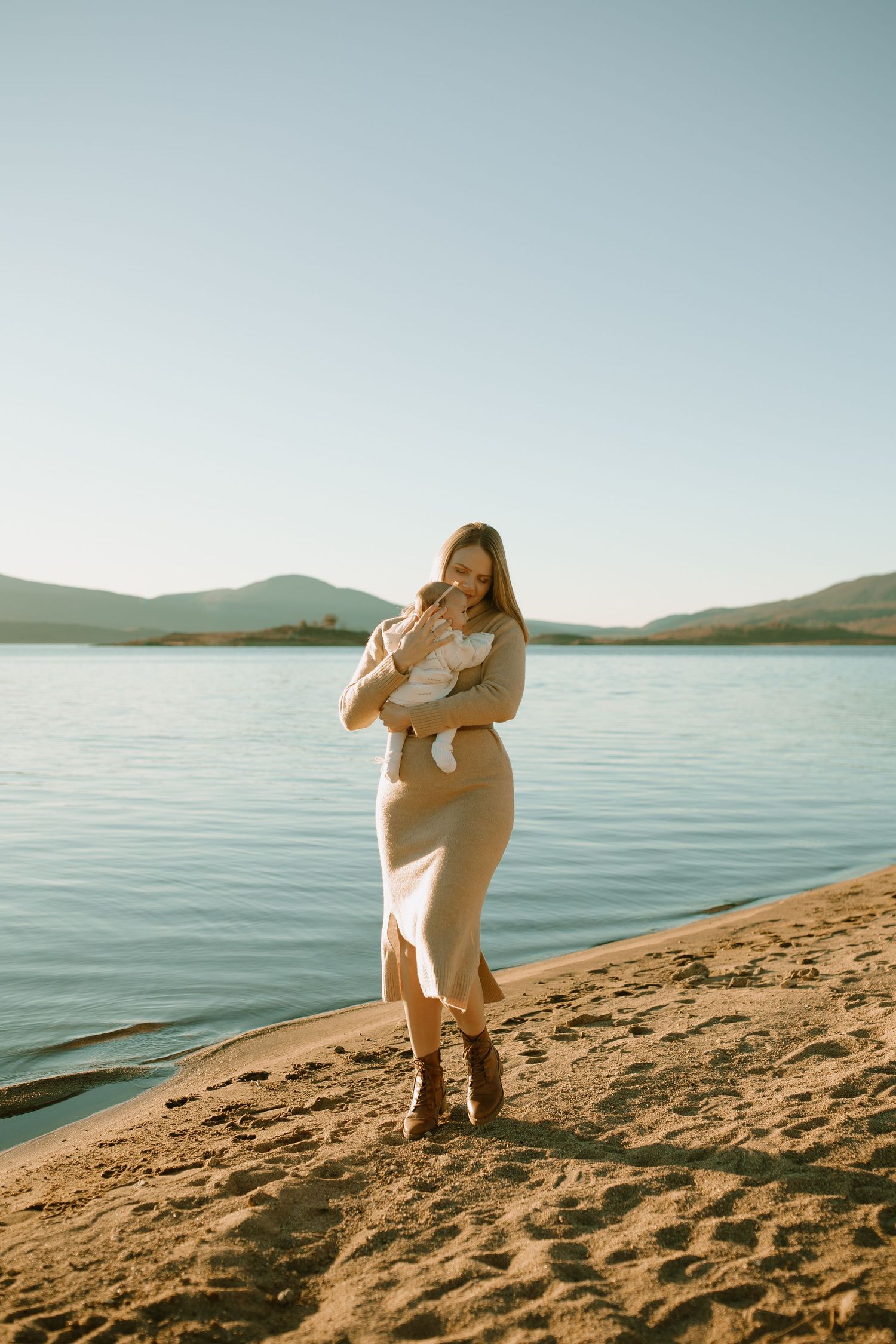Person in a beige dress holds a baby while standing on a sandy beach beside a calm lake with mountains in the distance.