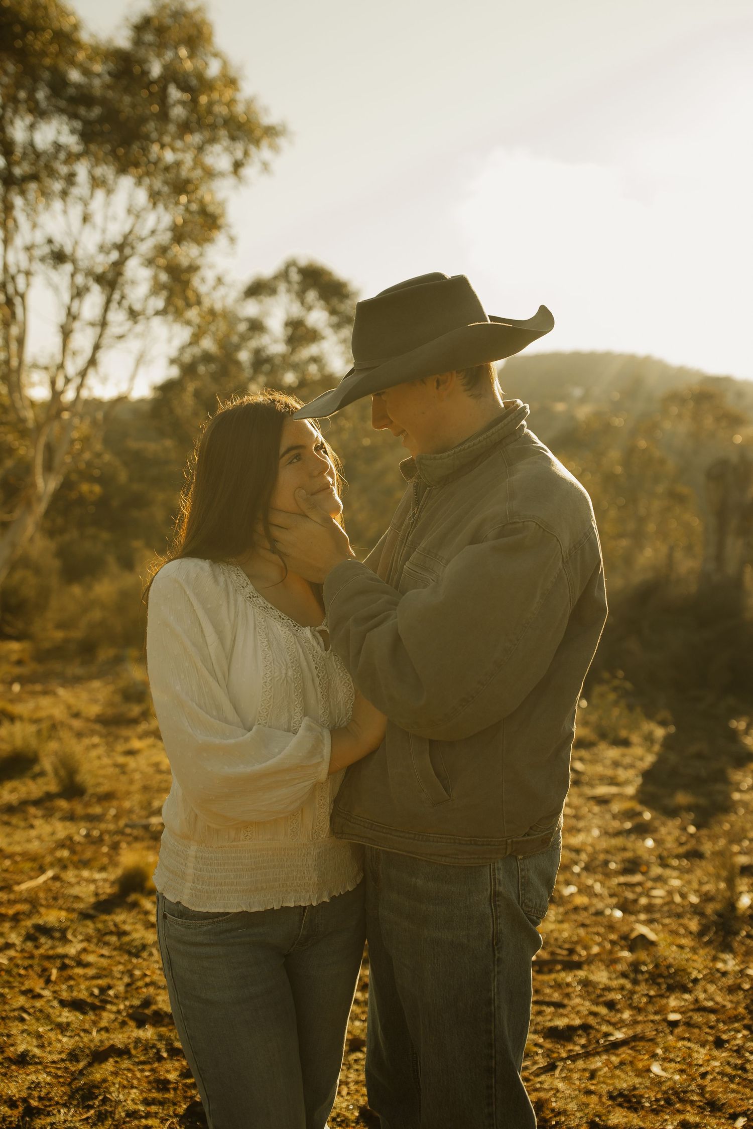 A romantic couple embraces in warm sunset light against a rural backdrop with eucalyptus trees.
