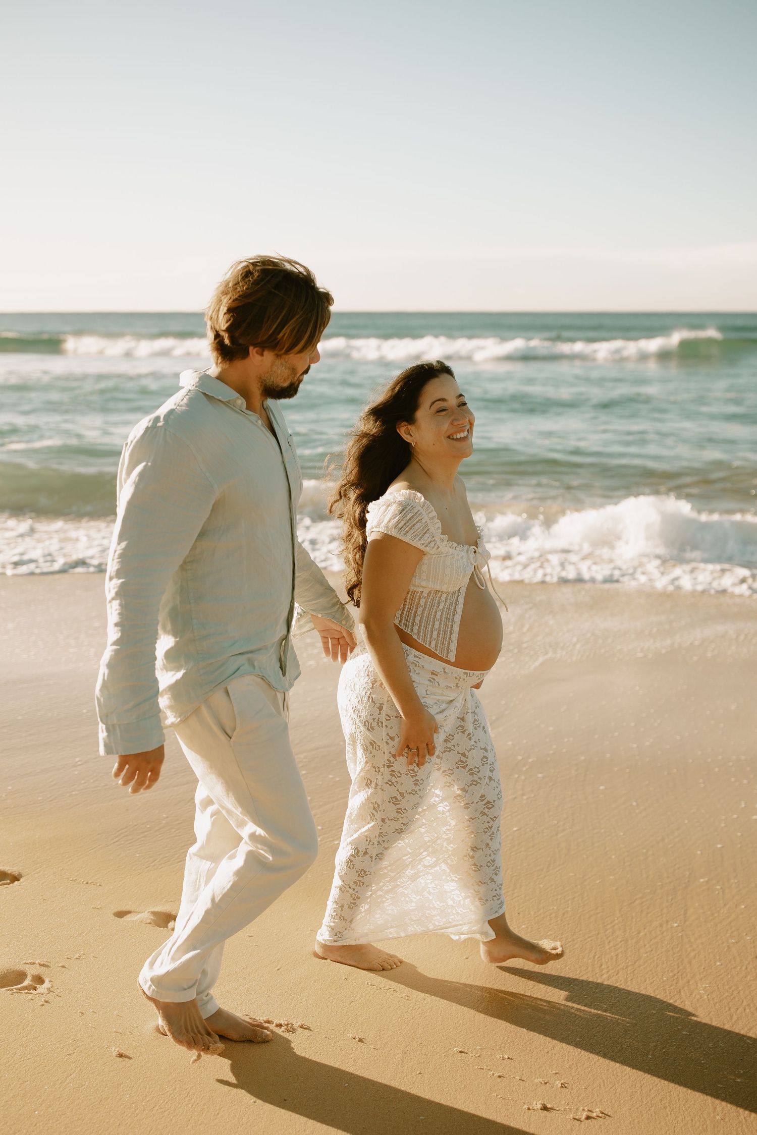 A couple walks together on a sandy beach with crashing waves, dressed in light colored clothing on a sunny day.