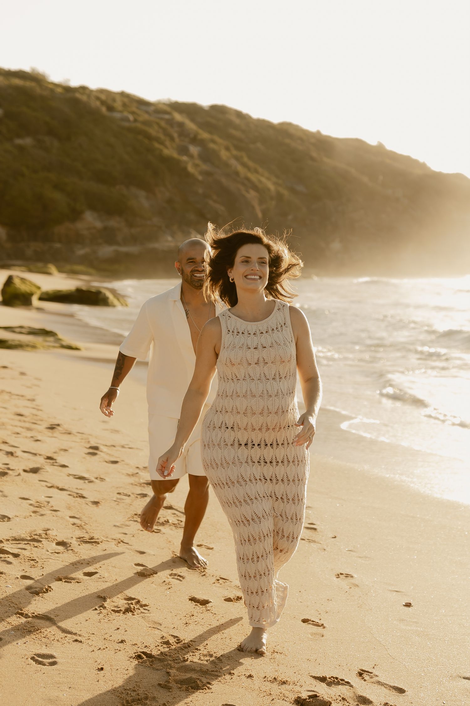Two people joyfully running along sandy beach at golden hour with waves crashing behind them.