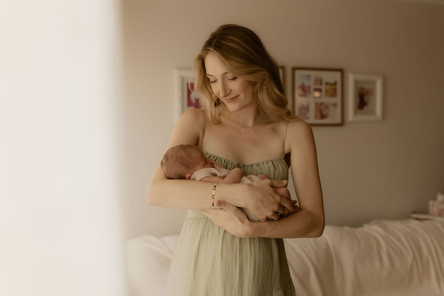 A woman in an olive green dress sits on a white bed in a softly lit room with framed artwork on the walls.