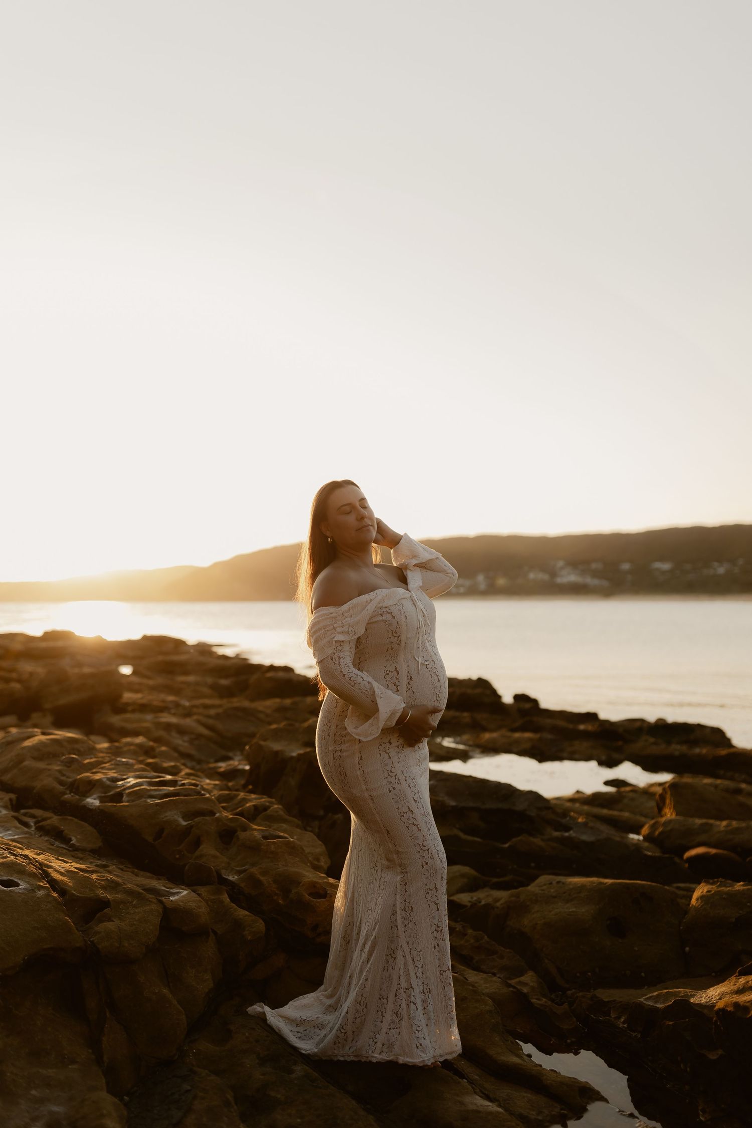 A couple embraces on rocky coastal cliffs during golden hour sunset by the ocean.
