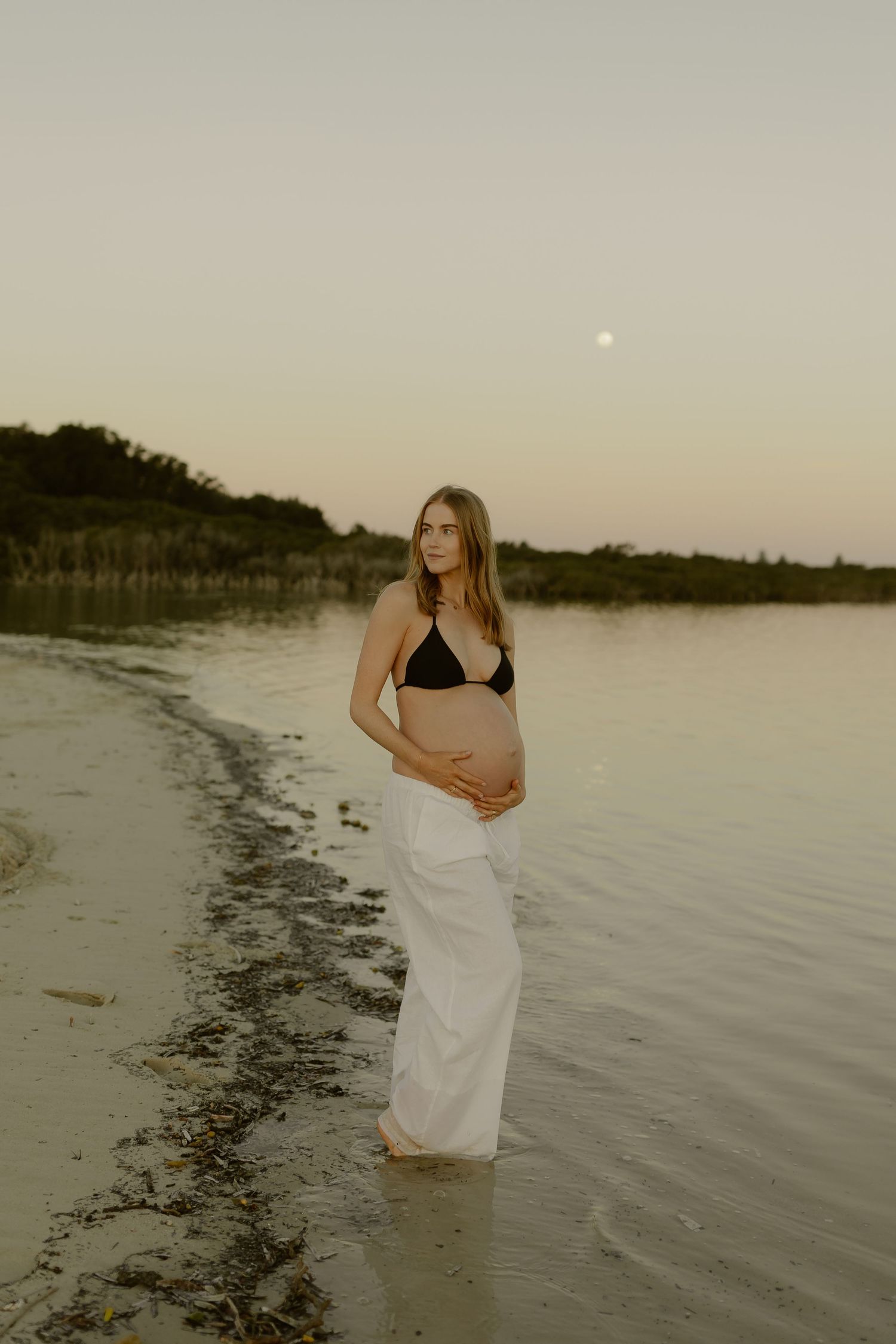 Pregnant woman in black bikini top and white skirt cradling belly on beach at sunset with moon visible.
