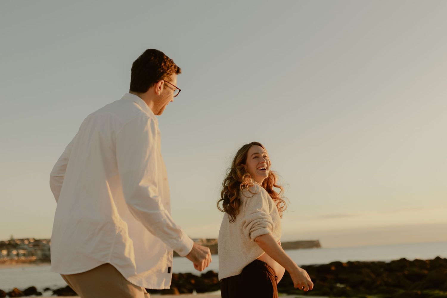 Couple holding hands laughing at golden hour sunset by rocky ocean shore, wearing white tops.