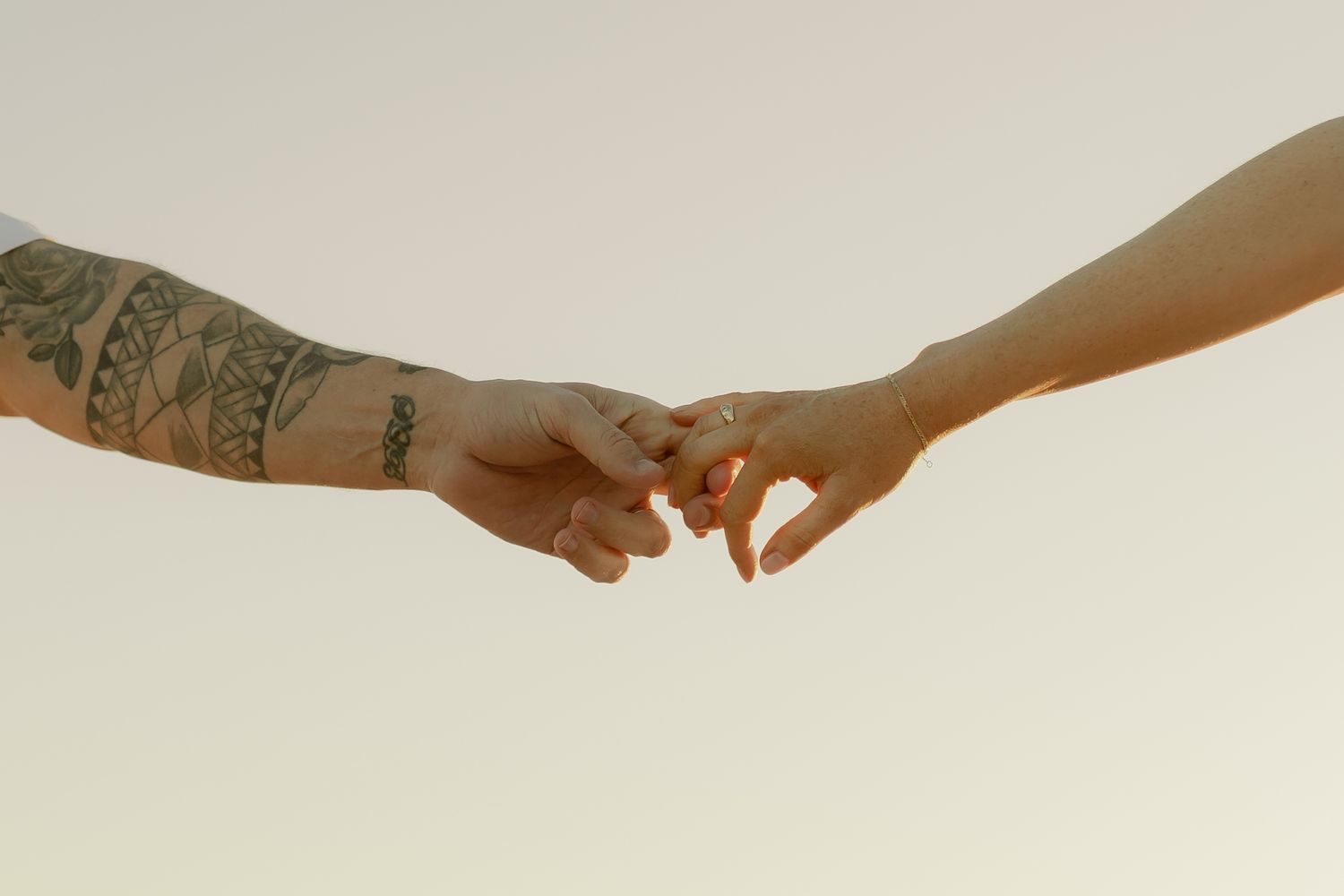 Two hands gently touching fingertips, one with tattoos, against a soft neutral background.