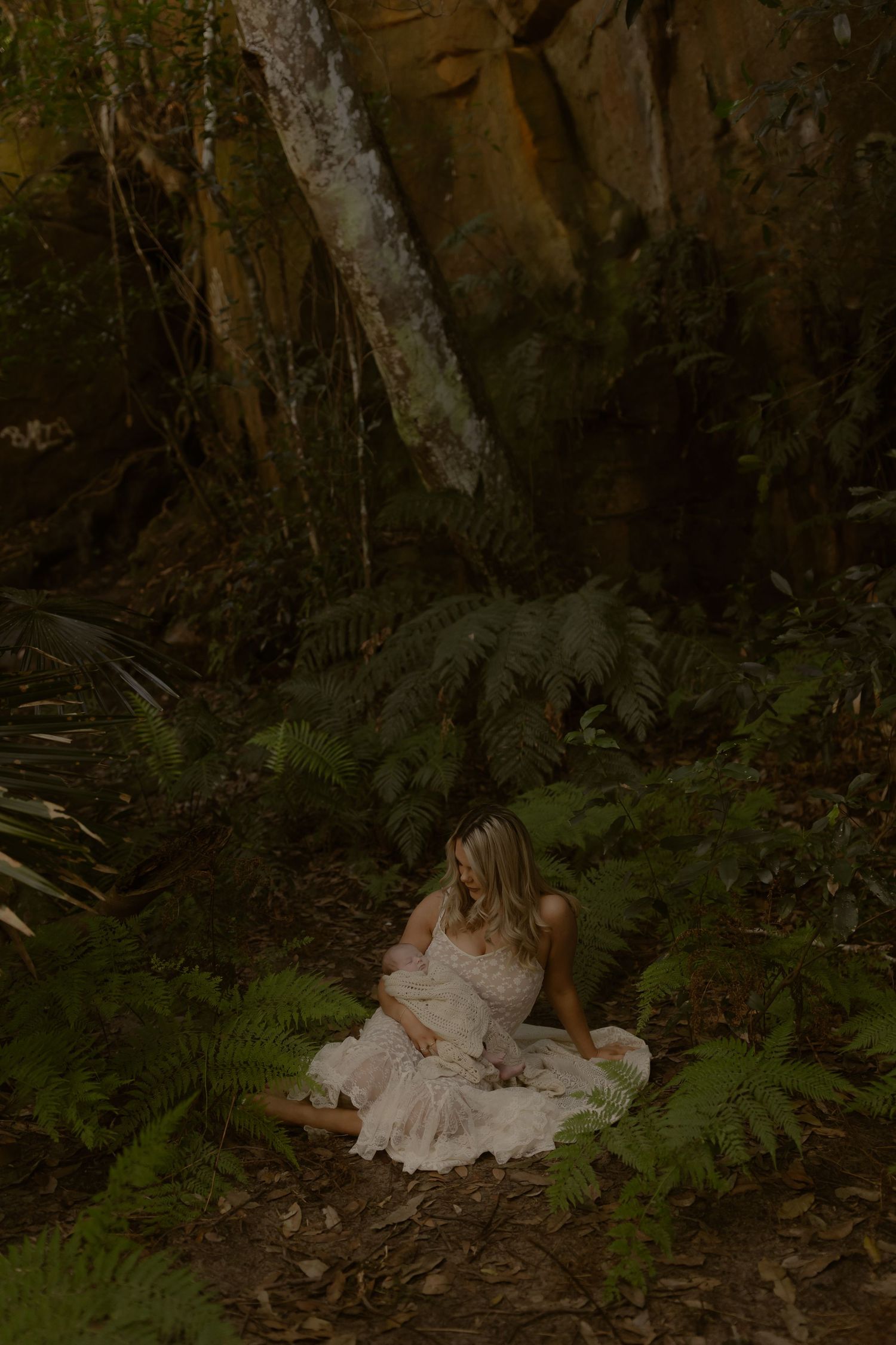 Pregnant woman in white lace dress sitting among ferns in a lush forest with rocky cliffs.