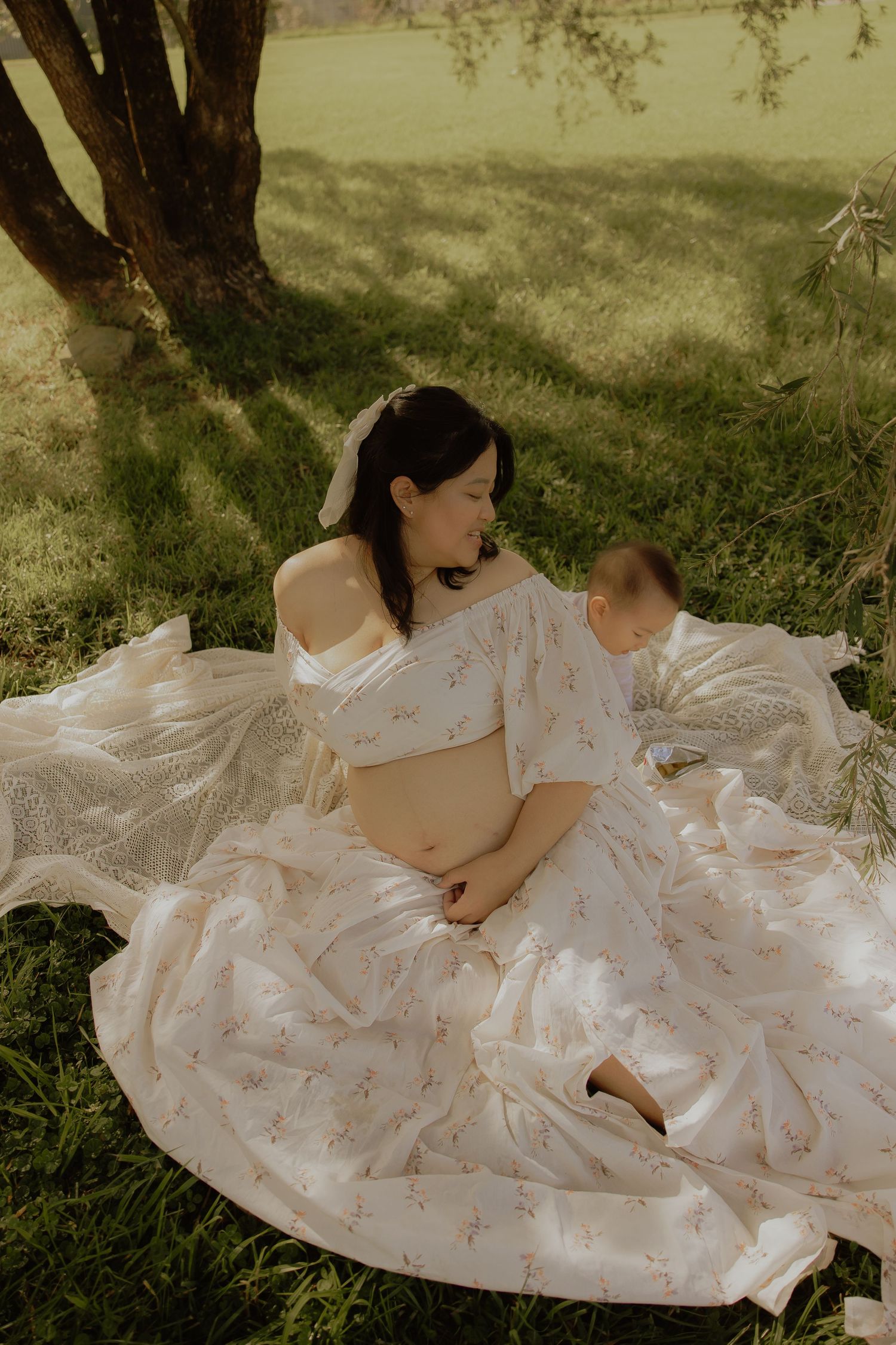 Pregnant woman in floral crop top and skirt sitting on grass with baby during outdoor maternity photoshoot.