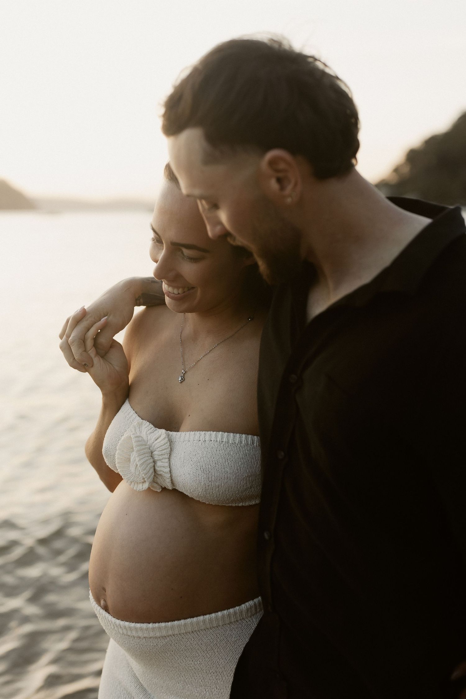 An intimate waterfront maternity photo of a couple sharing a tender moment at sunset.