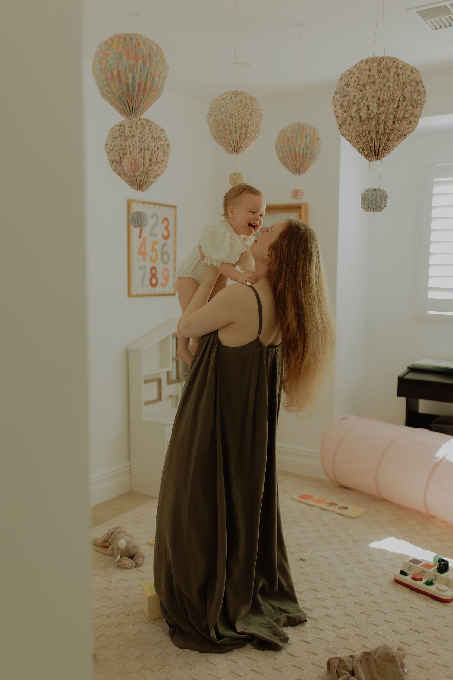A mother in an olive dress lifts her laughing baby in a decorated nursery room.