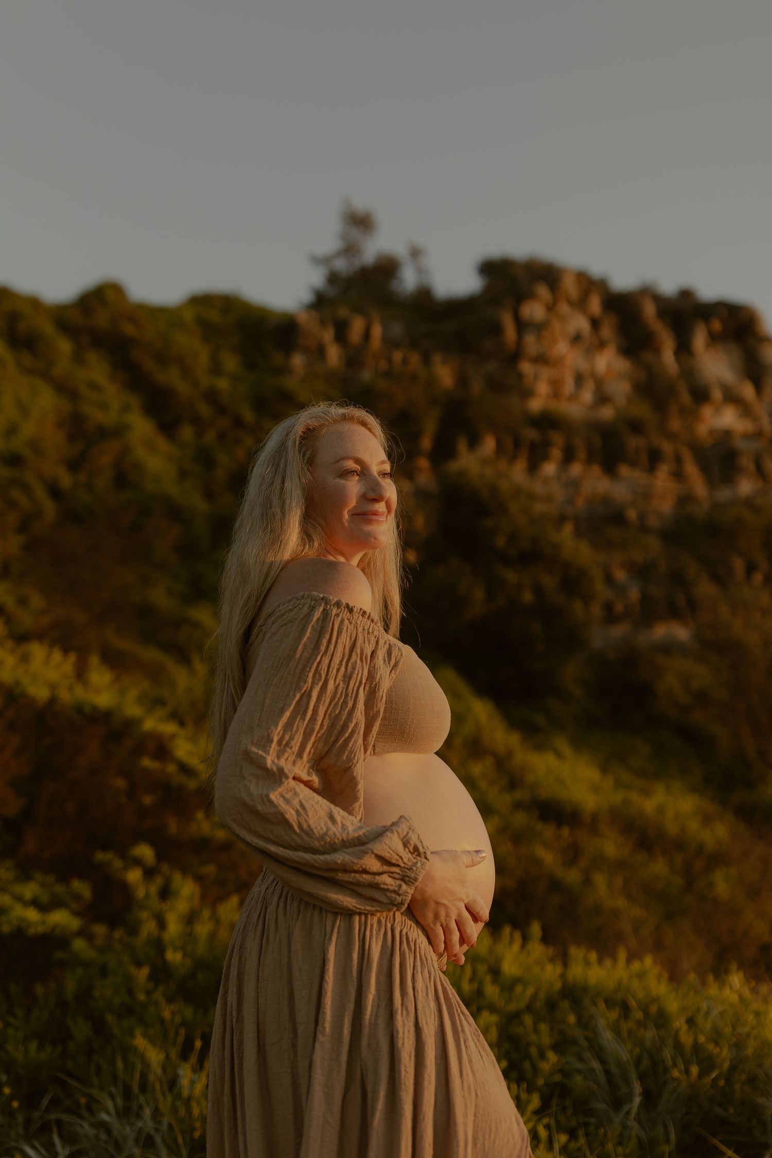 Smiling pregnant woman in off-shoulder beige dress cradling her belly outdoors at sunset with rocky cliffs behind her.